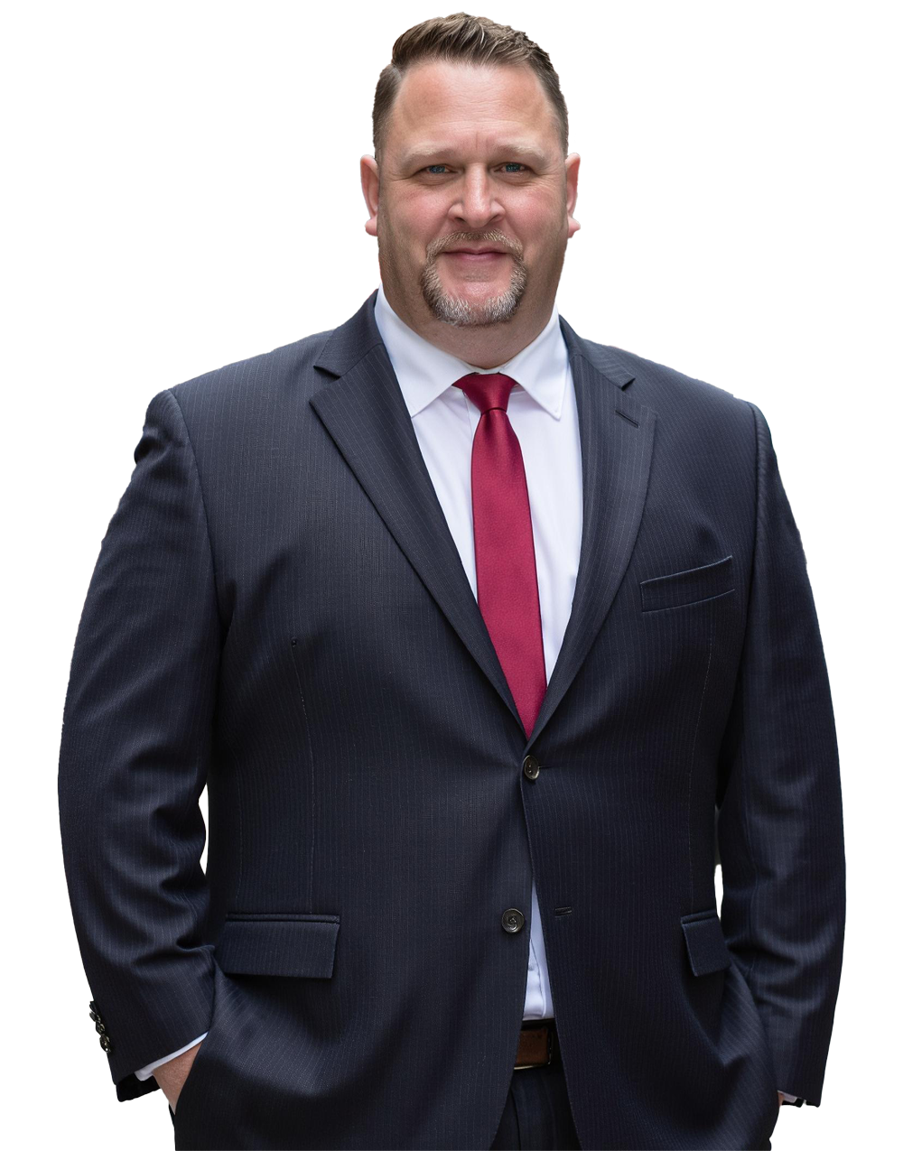 A man in a dark blue suit, white shirt, and red tie posing confidently with hands in his pockets, against a plain background.