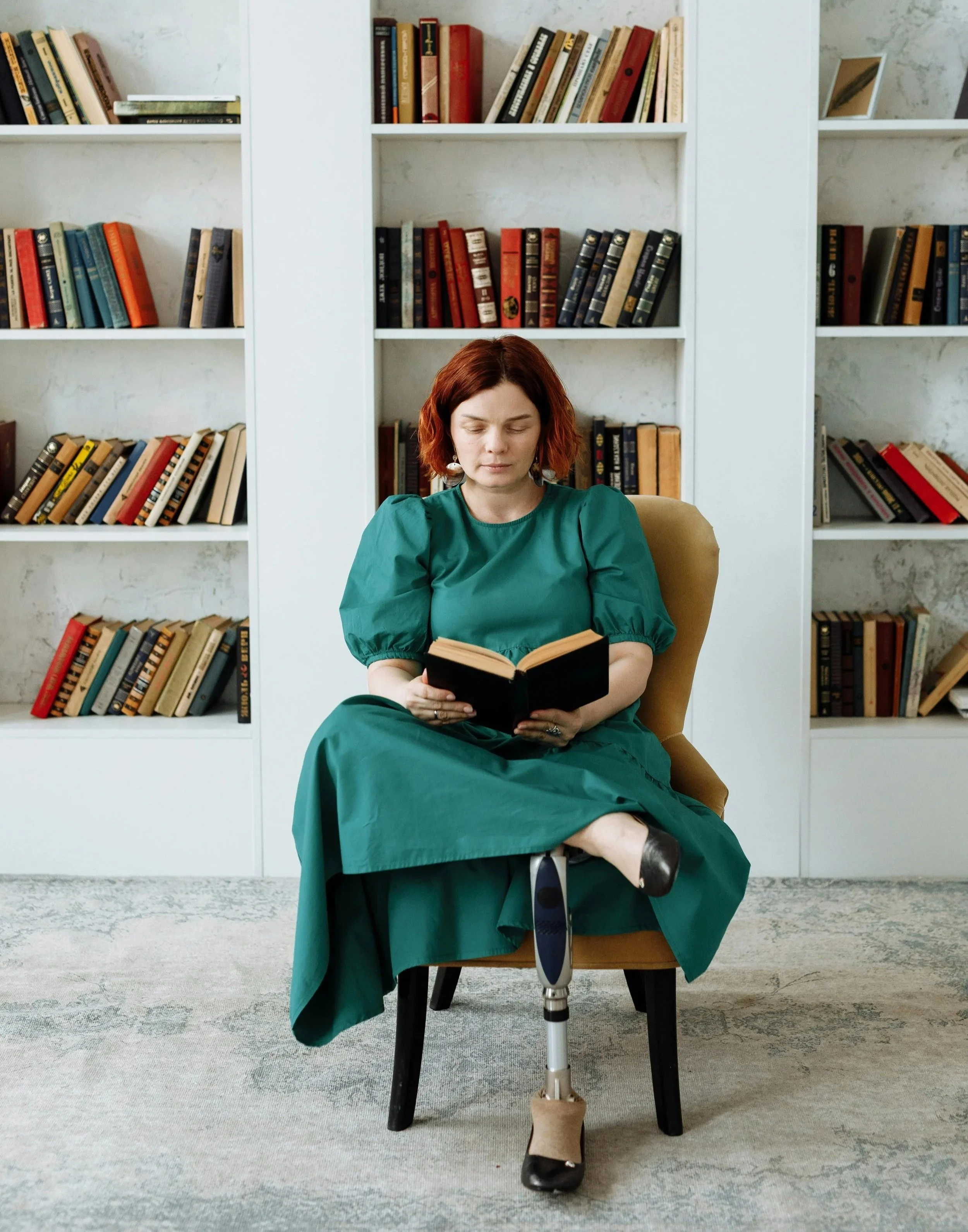 Young Lady sitting Reading a Book in her home.  She wears a green dress.  She has a right below the knee amputation and has a prosthetic leg.