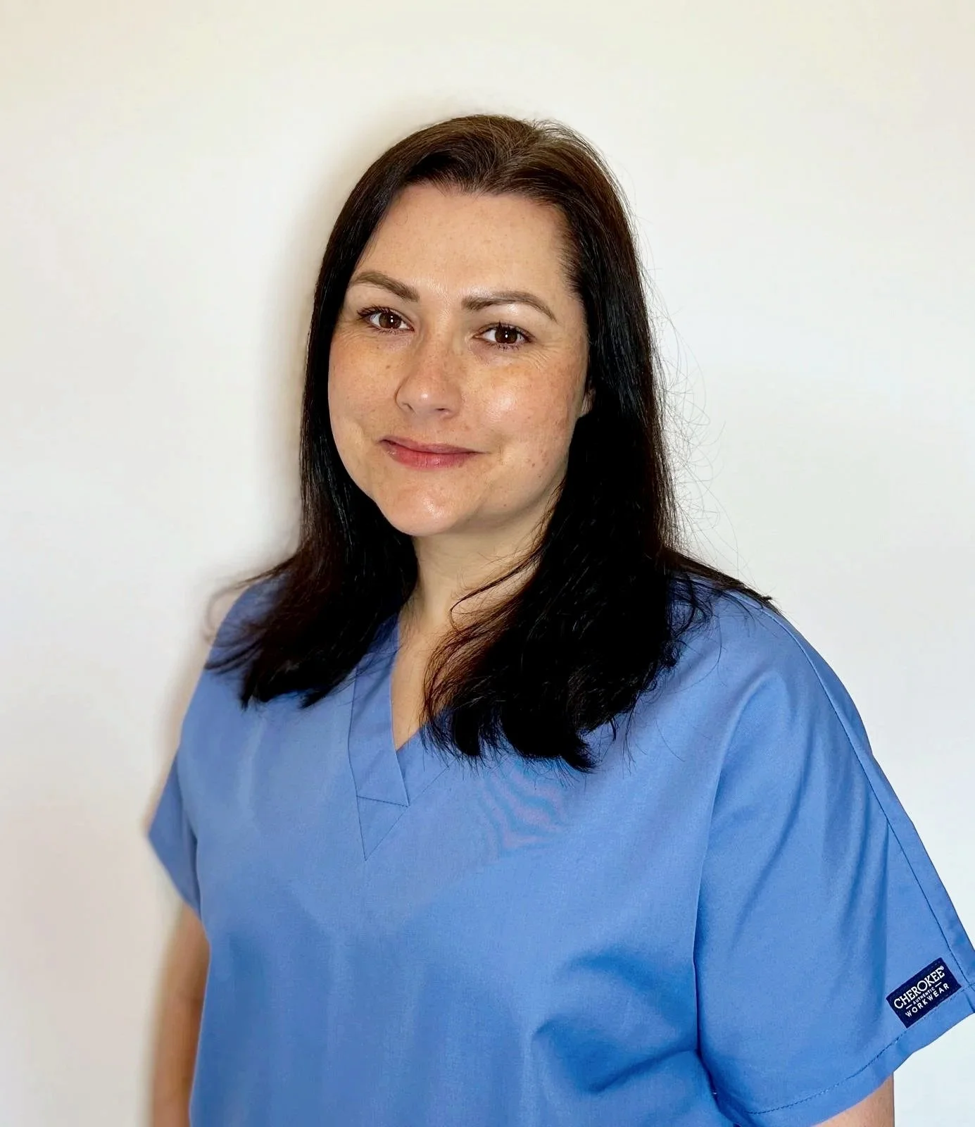 A woman with shoulder-length dark brown hair, wearing light blue medical scrubs, standing against a plain white background. She is smiling slightly and looking directly at the camera.