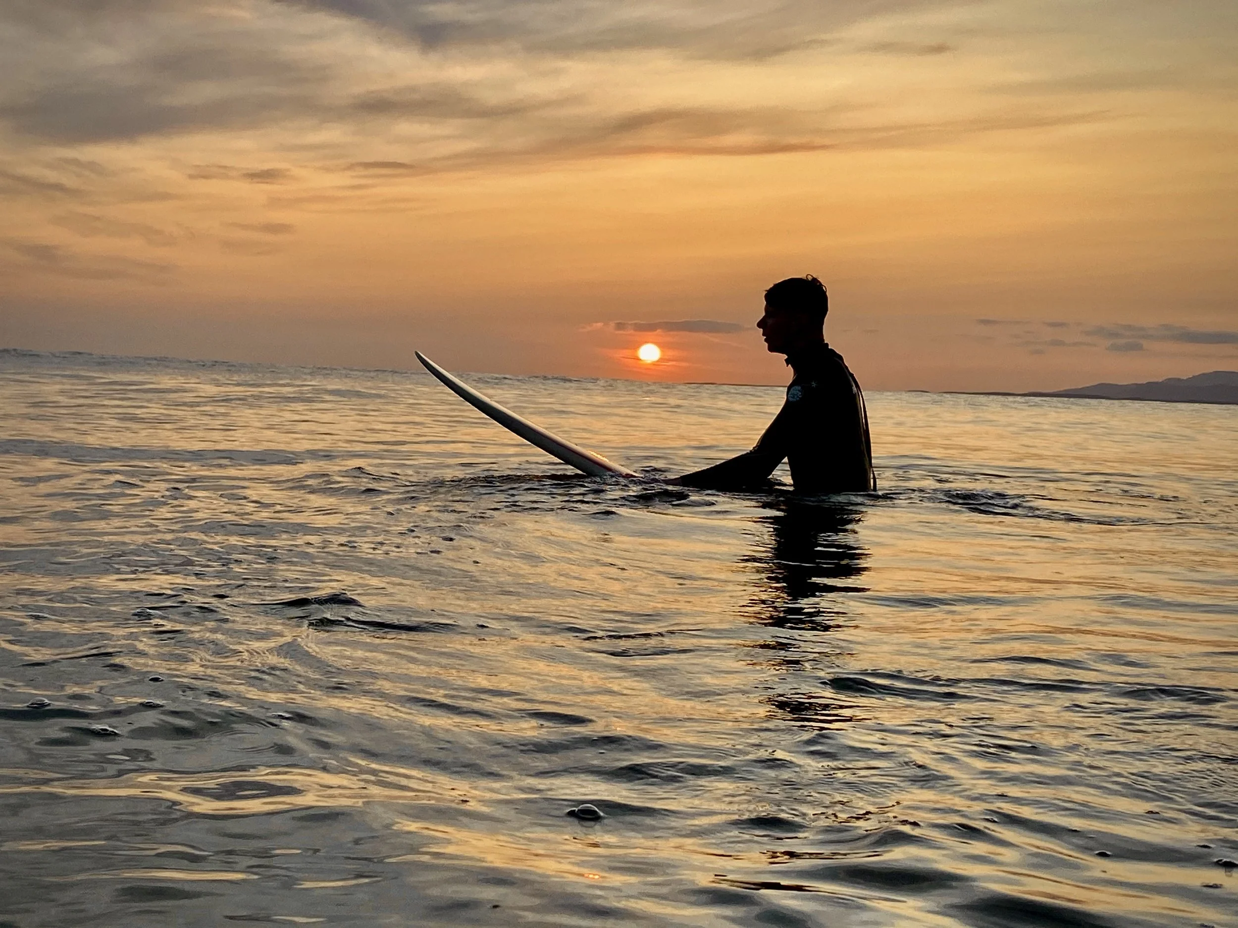 A person holding a surfboard while sitting in the water during sunset, with a colorful sky and distant mountains in the background.