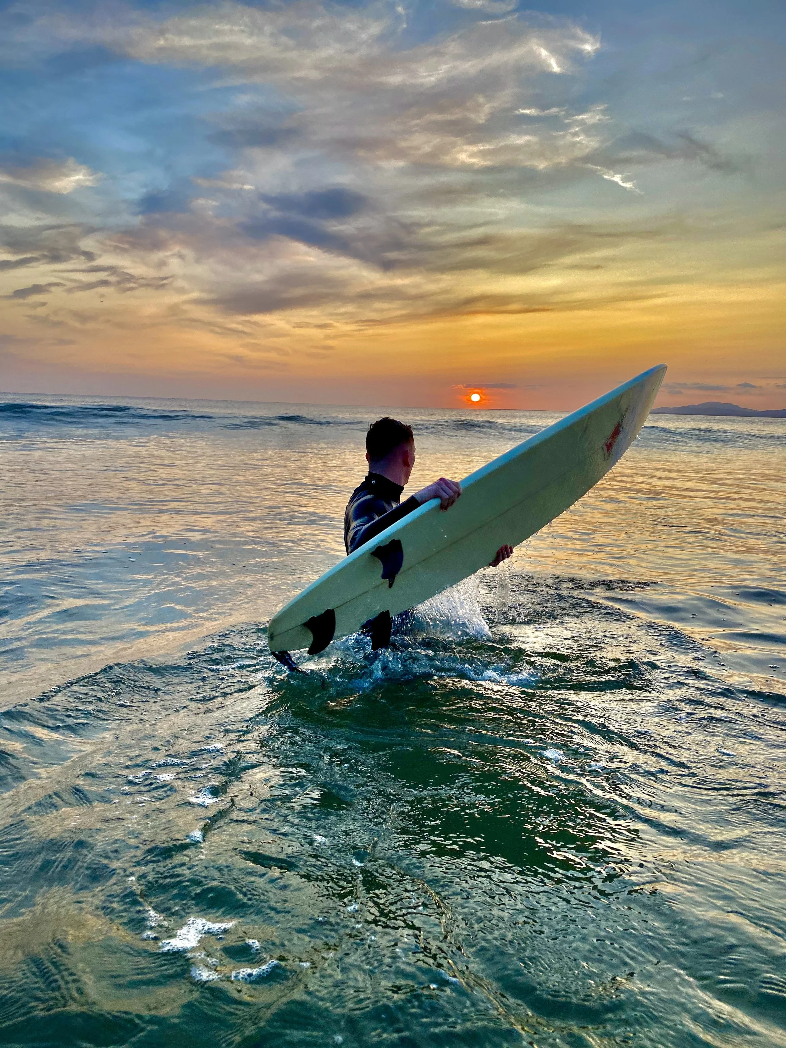 Person holding a surfboard while walking in the ocean at sunset.