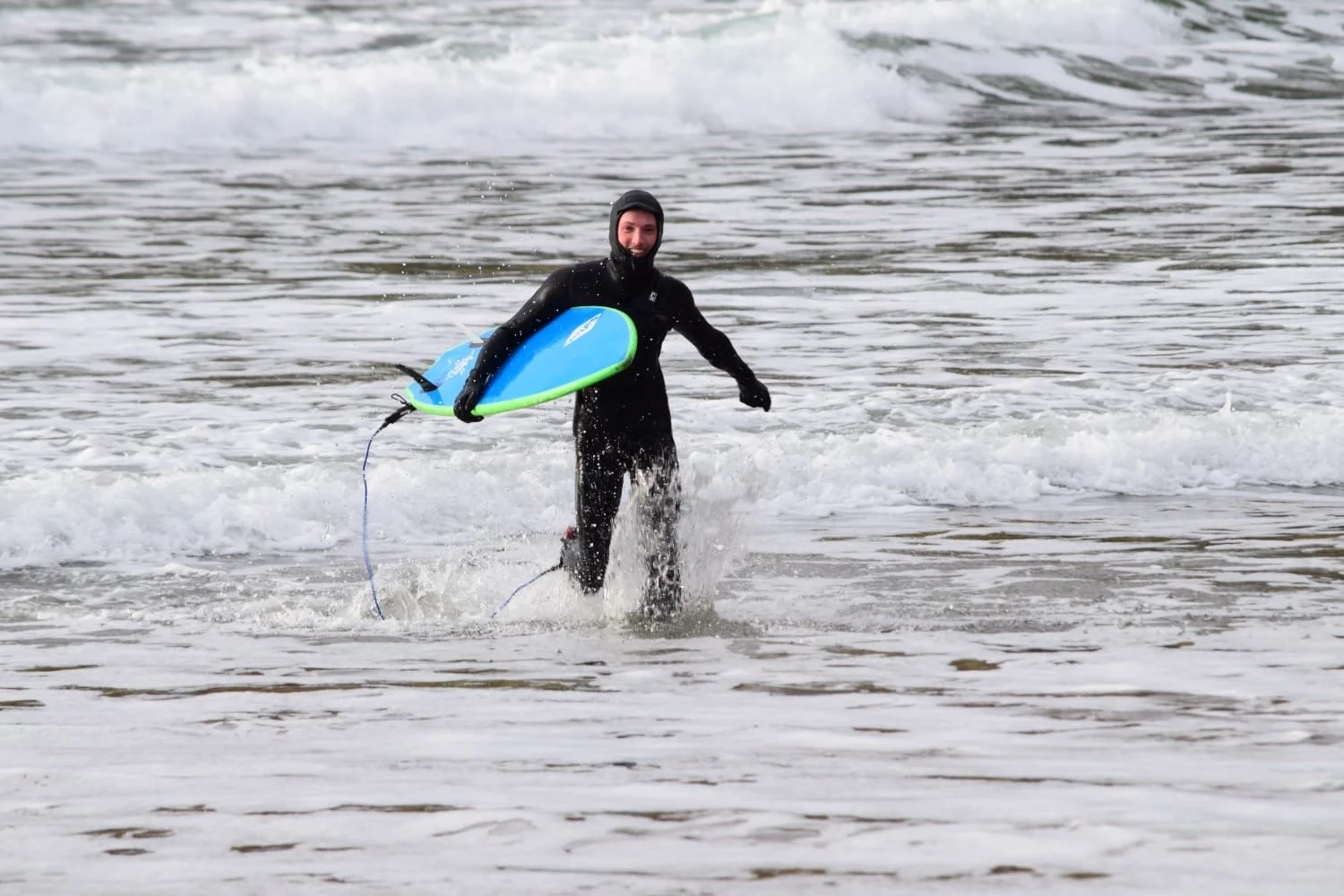 A person in a black wetsuit and hood holding a blue and green surfboard while walking into the ocean.