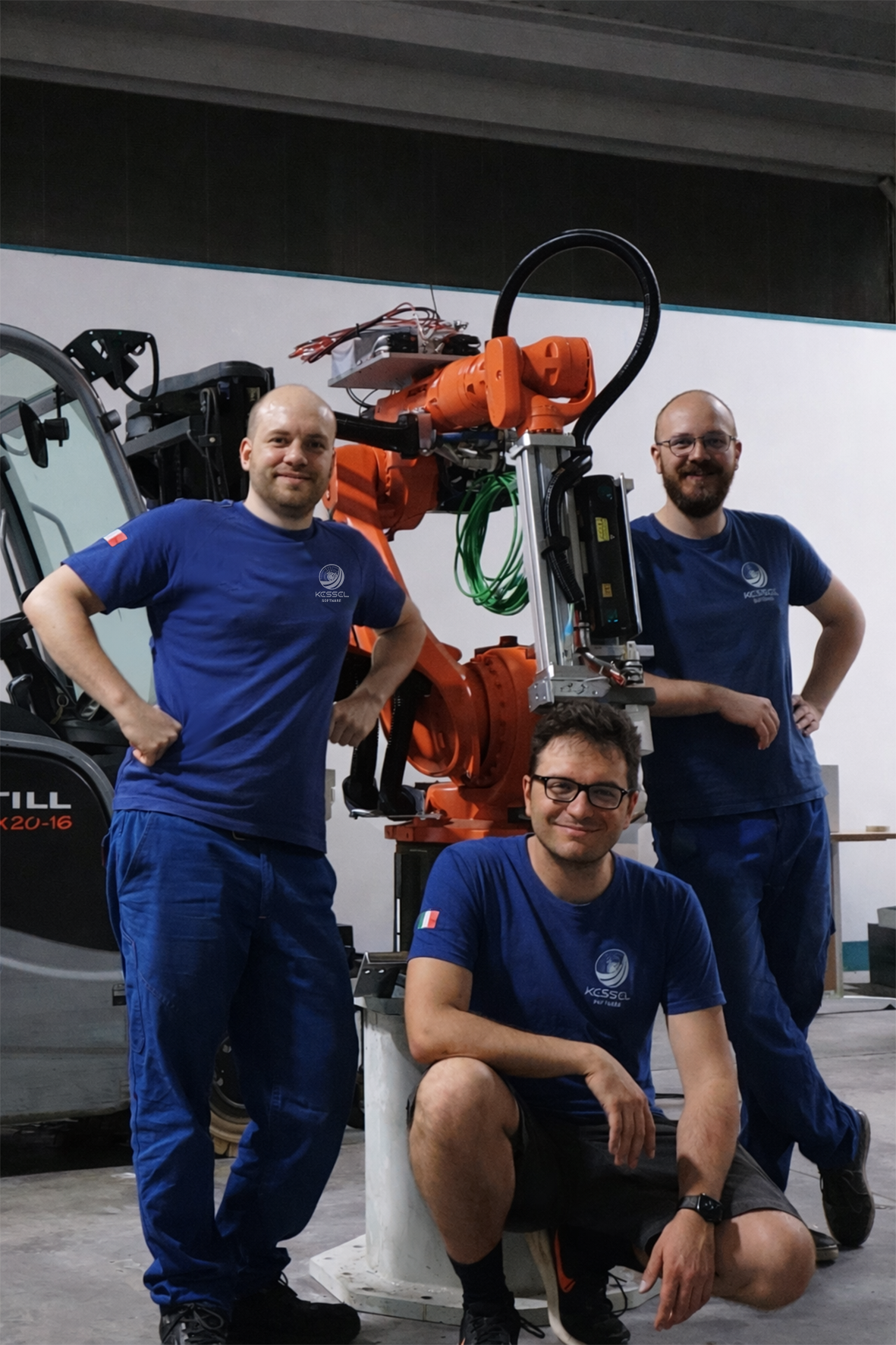 Tre persone con magliette blu e logo di un'organizzazione scientifica, una di loro seduta e due in piedi, in un laboratorio con un robot industriale arancione.