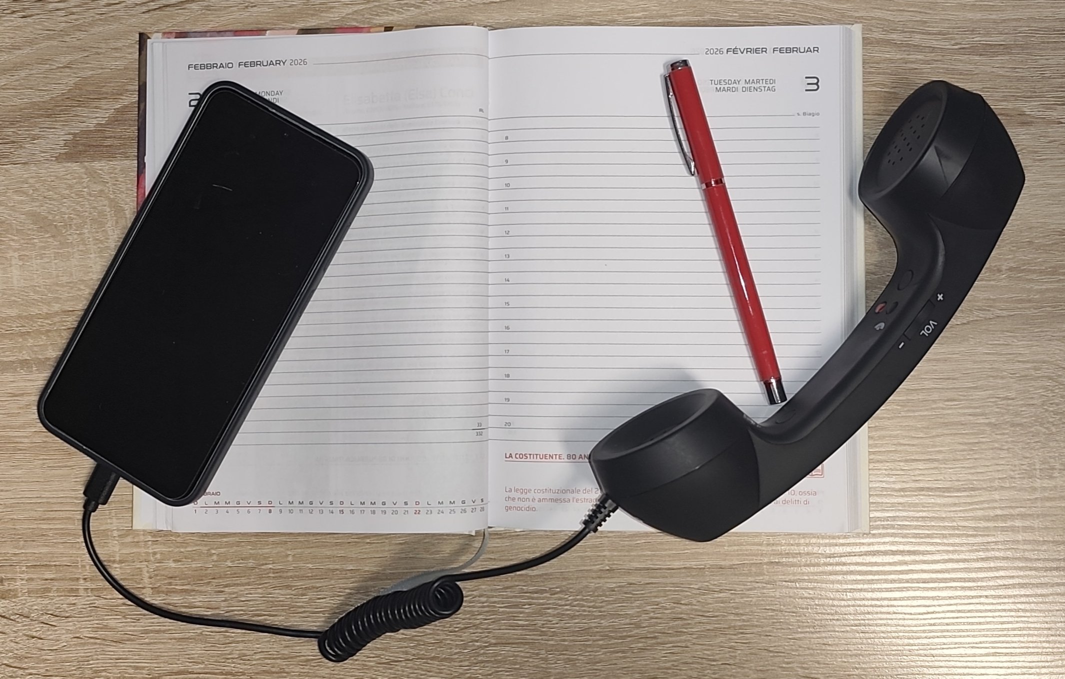Un pianificatore aperto su un tavolo di legno con una penna rossa, un telefono cellulare nero con il cavo di alimentazione e un telefono fisso scuro collegato con filo.