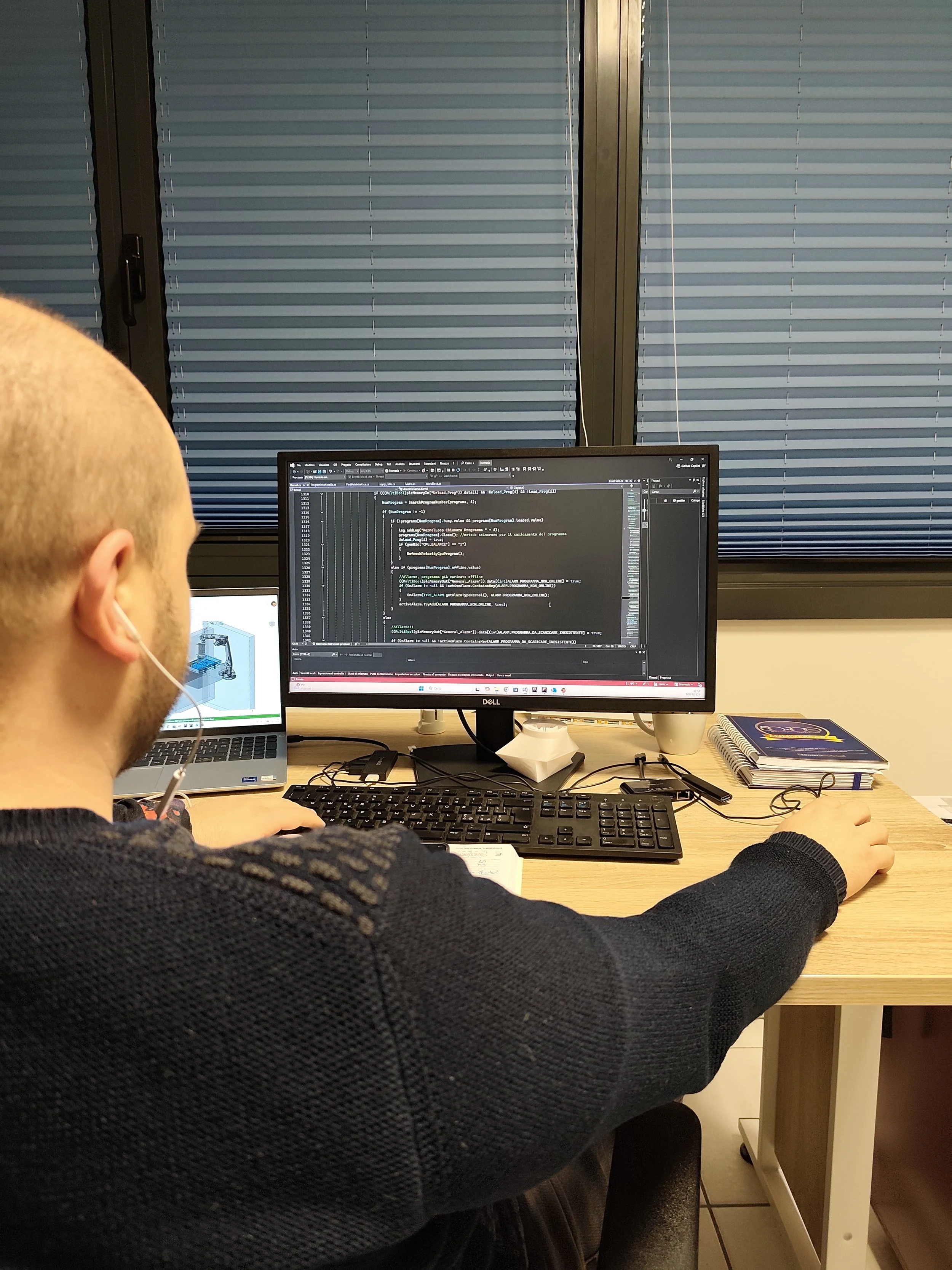 Uomo che lavora davanti a schermo di computer con codice di programmazione, in un ufficio con finestre con tapparelle abbassate.