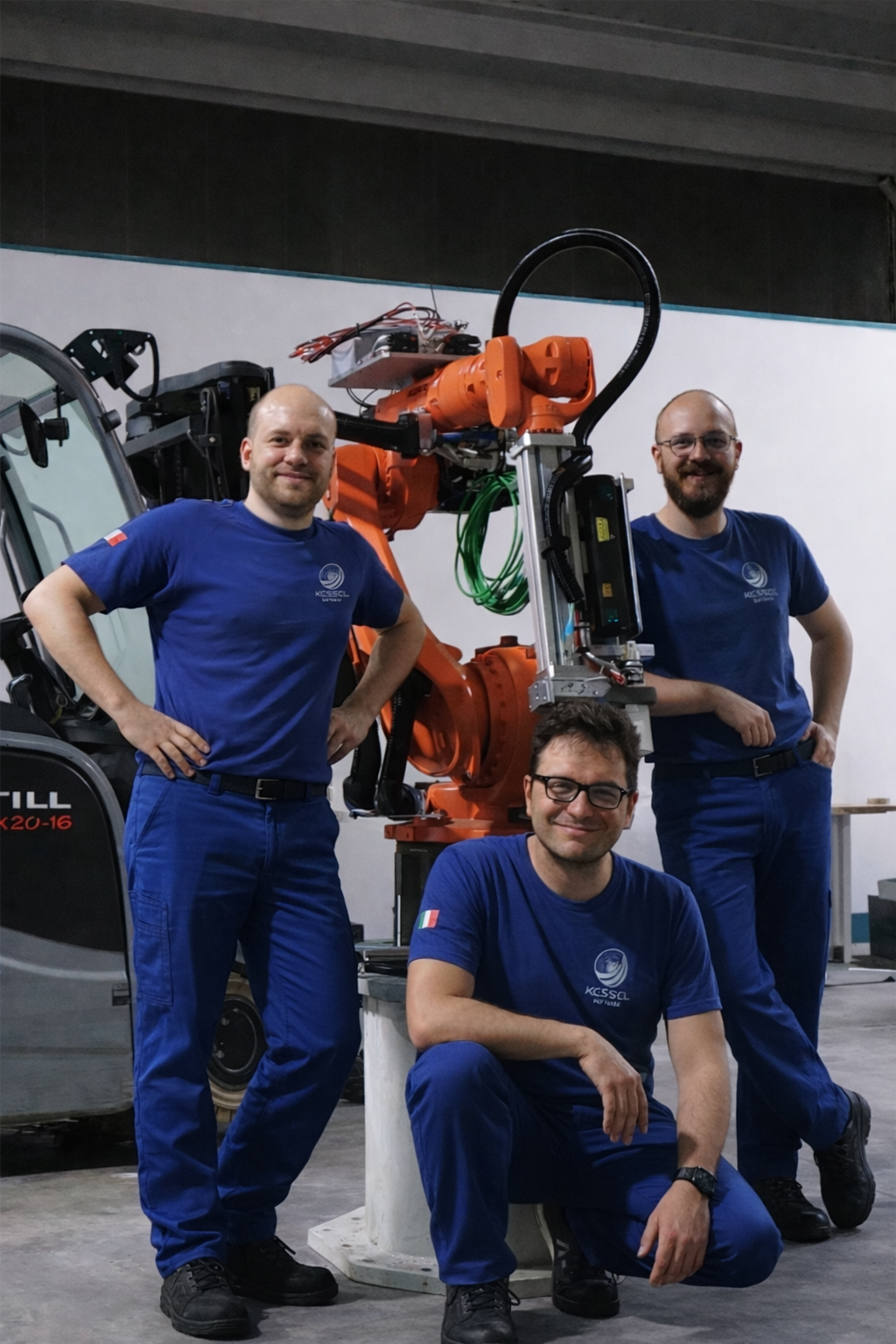 Tre uomini con abbigliamento blu posano accanto a un robot industriale arancione e a un veicolo simile a un carro agricolo all'interno di un laboratorio o officina.