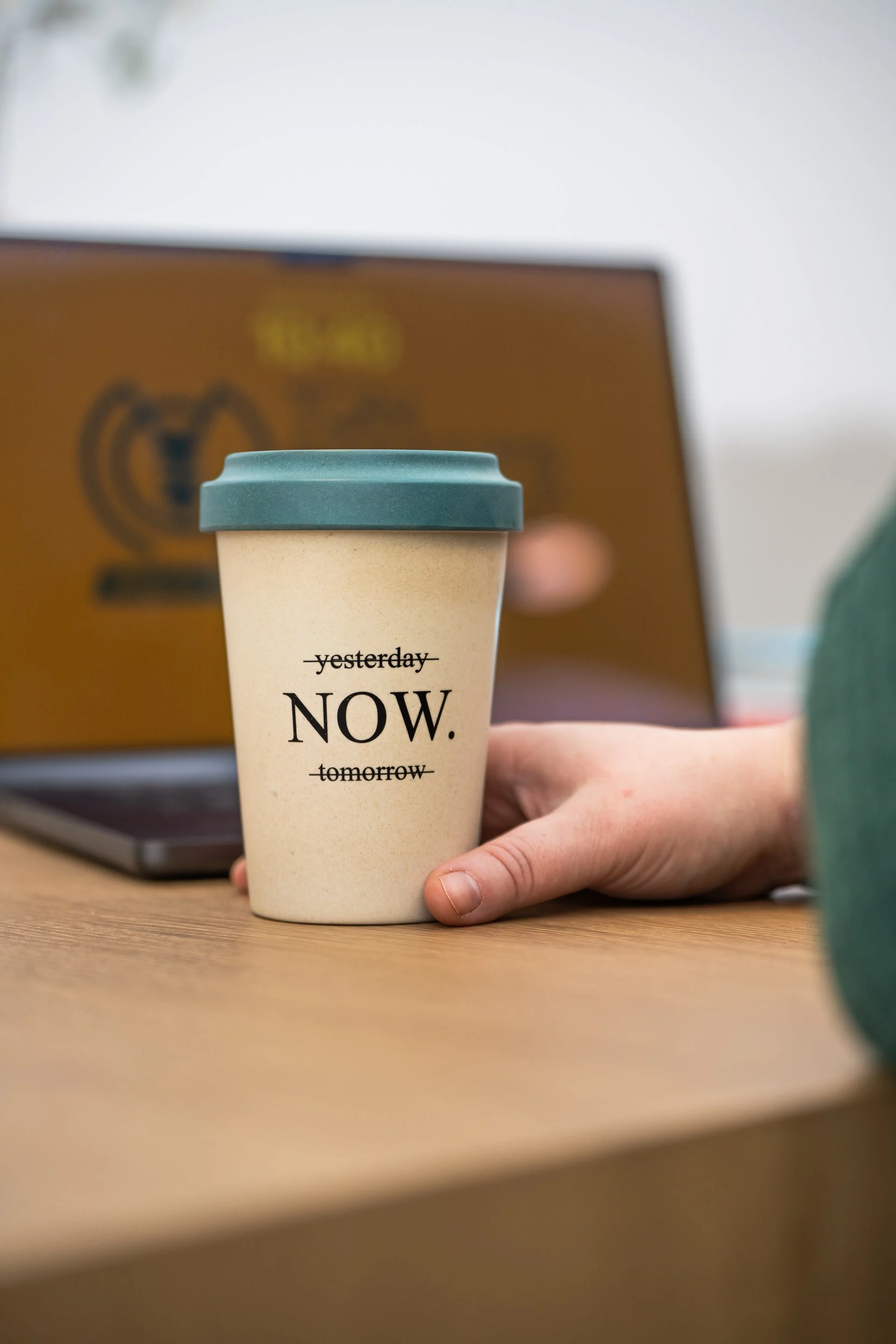 Une tasse en papier beige avec un couvercle bleu, tenue par une main humaine, sur un bureau en bois, avec un arrière-plan flou comprenant une boîte en carton.