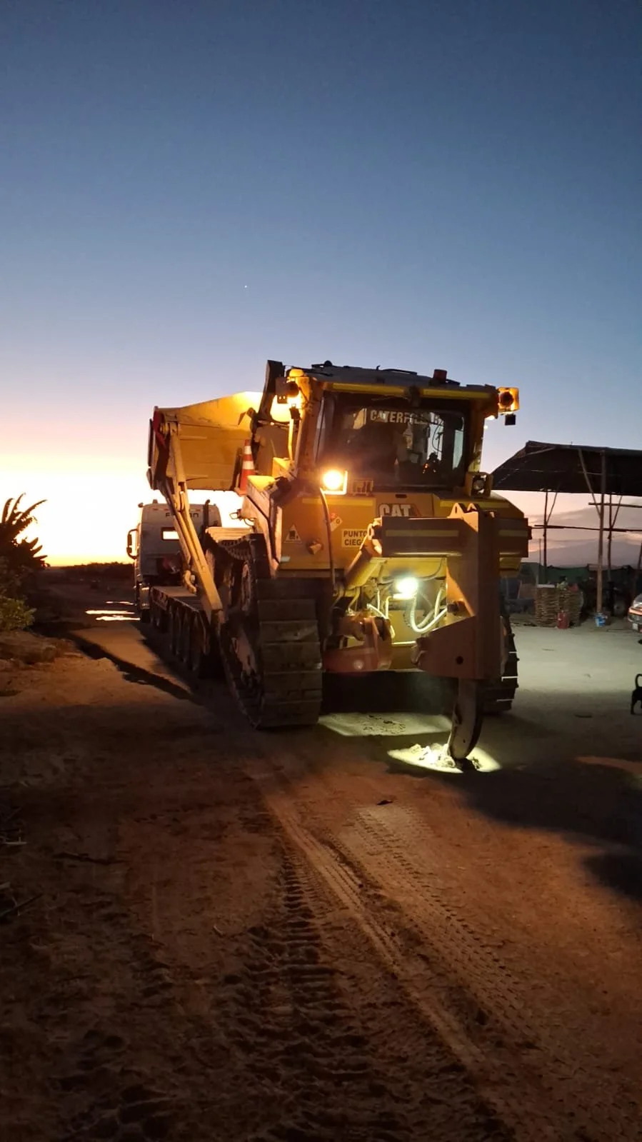 Una maquinaria de construcción en medio de un camino de tierra al atardecer, con luces encendidas y un cielo en tonos azul y naranja en el fondo.