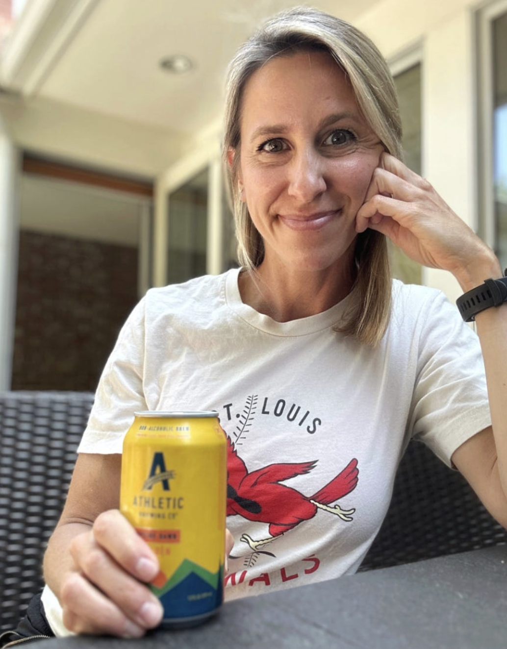A woman sitting outdoors holding a yellow can of Athletic Beer, smiling at the camera, wearing a white T-shirt with a red bird graphic and the words 'St. Louis Wildcats', and wearing a black wristwatch.