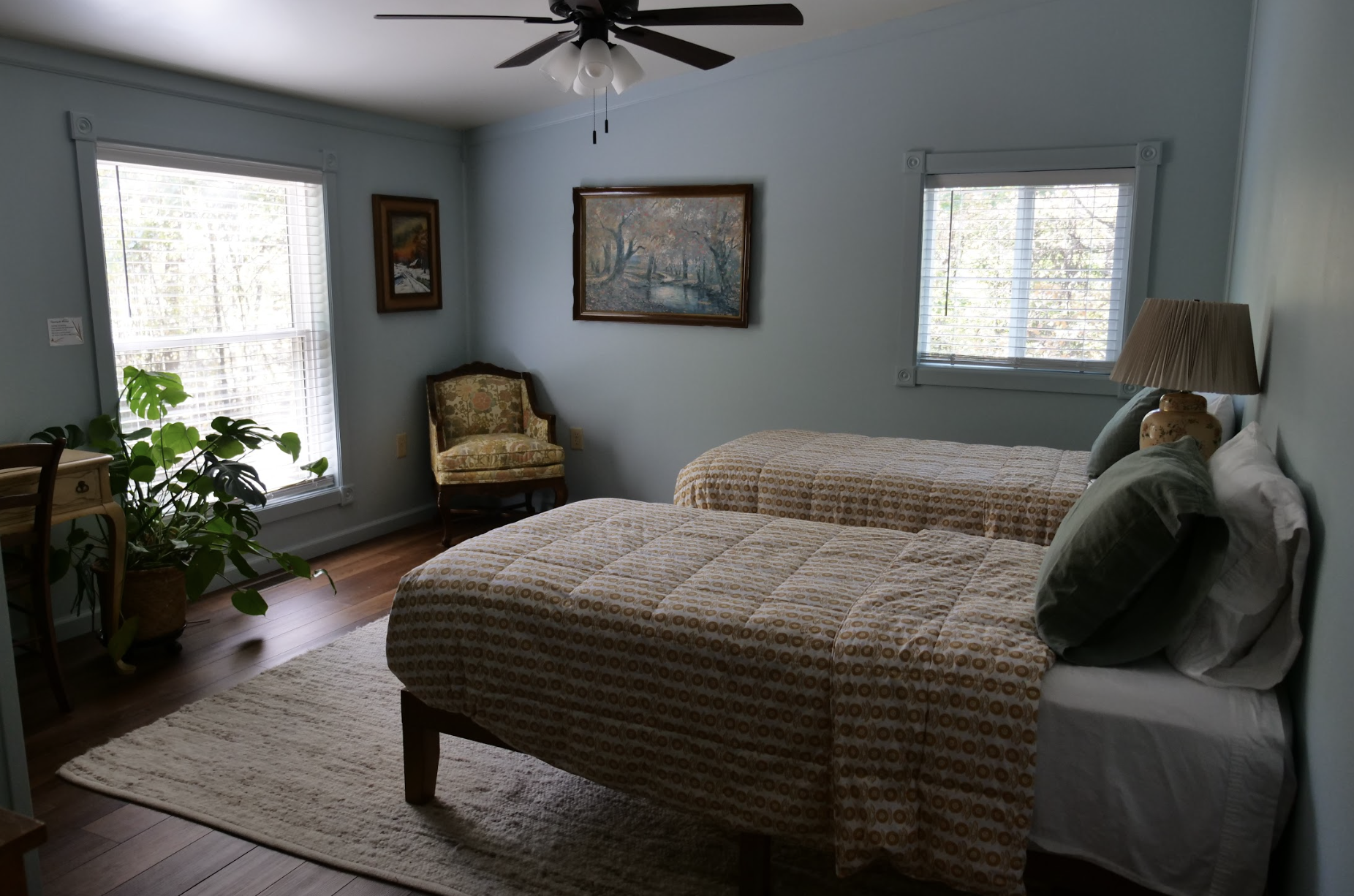 A cozy bedroom with two single beds, patterned bedspreads, a table lamp, and artwork on light blue walls. Contains a window with blinds, a decorative chair, and a large plant near the window.