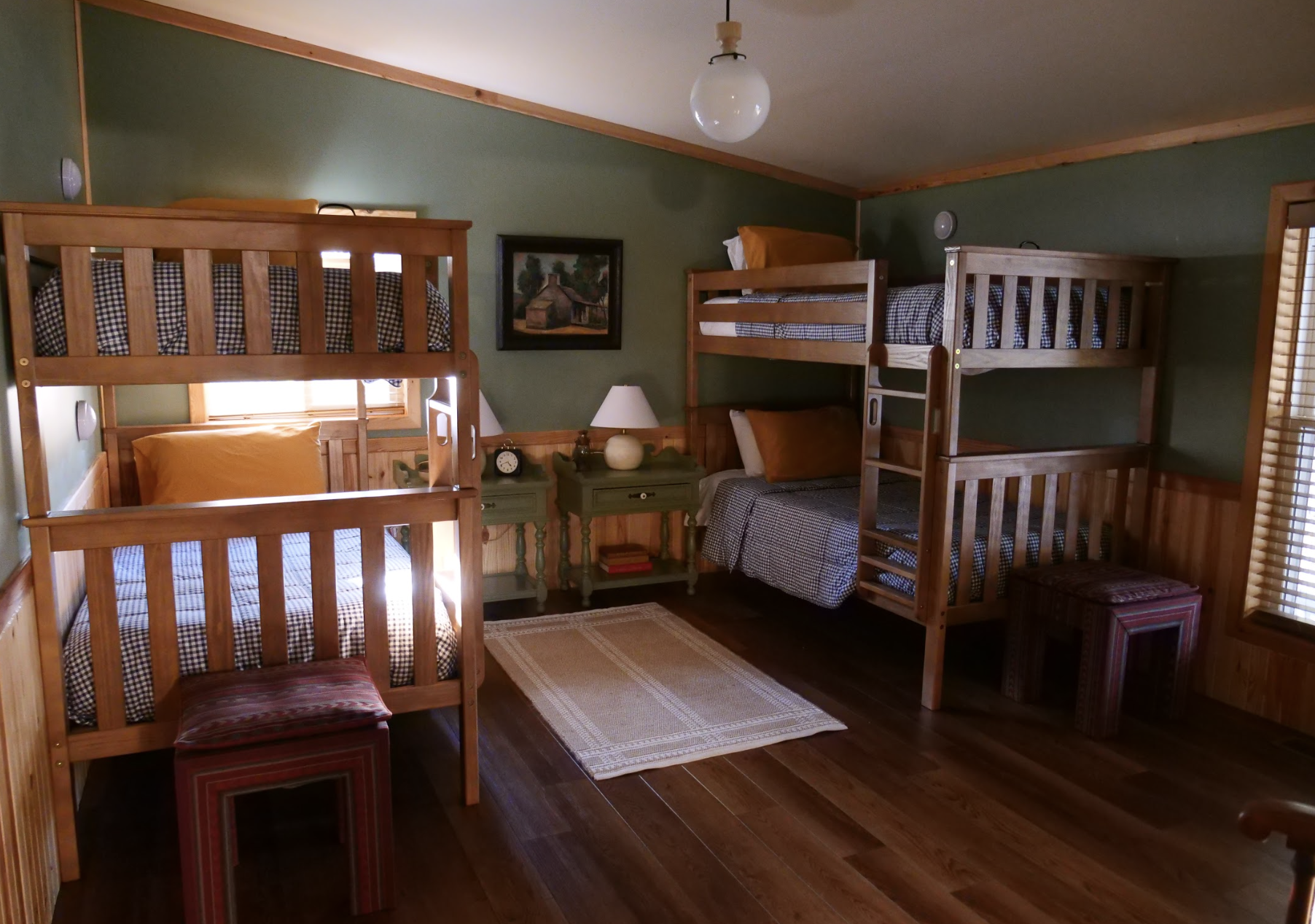 A rustic bedroom with four wooden bunk beds, three on the right and one on the left, each with plaid bedding. A window behind the left bunk beds lets in natural light. A small, dark green nightstand with a white lamp, clock, and books is between the beds against a green wall with wooden paneling at the bottom. A framed painting of a house hangs above the nightstand. A beige rug covers part of the wooden floor, and a small red plaid stool is in the corner near the right side of the room.