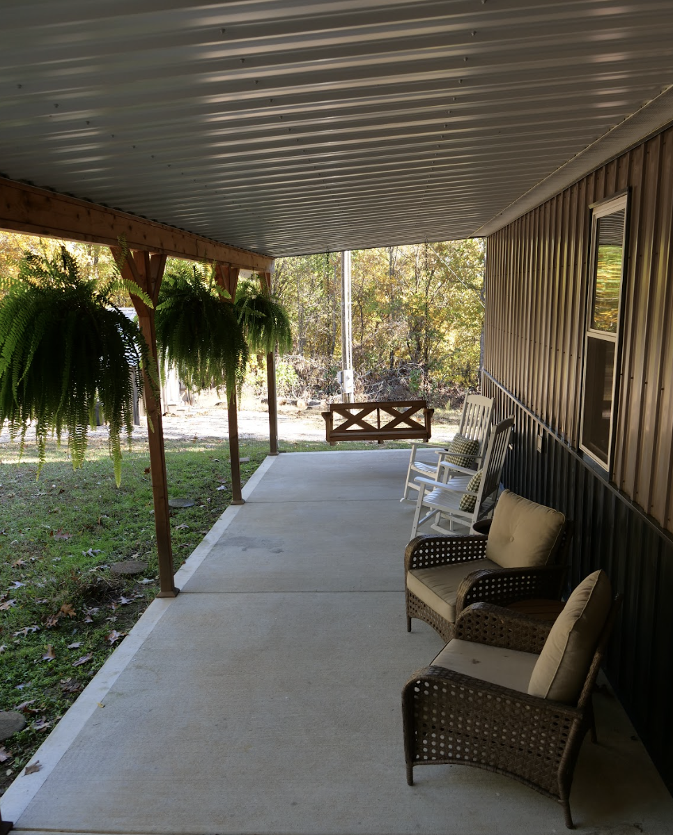 Front porch with hanging ferns, rocking chairs, and cushioned wicker chairs.