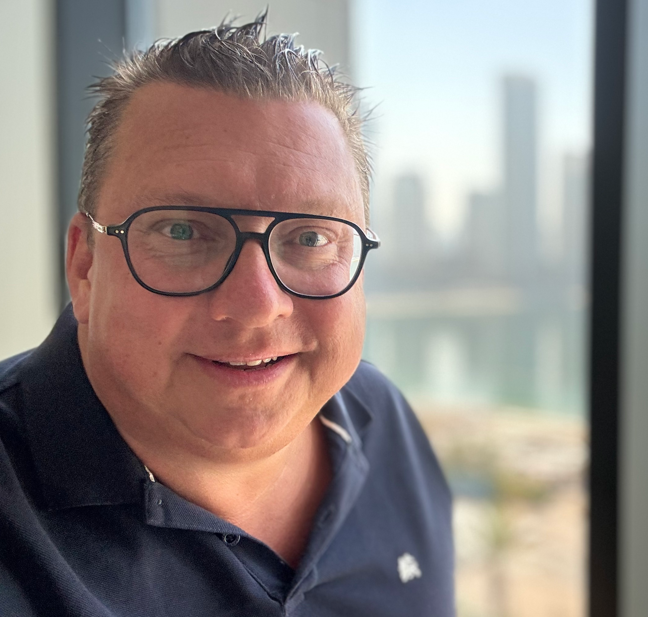 Nahaufnahme eines lachenden Mannes mit dunklen Haaren, Brille und dunklem Polo vor einem Fenster mit Stadtblick bei Tageslicht.