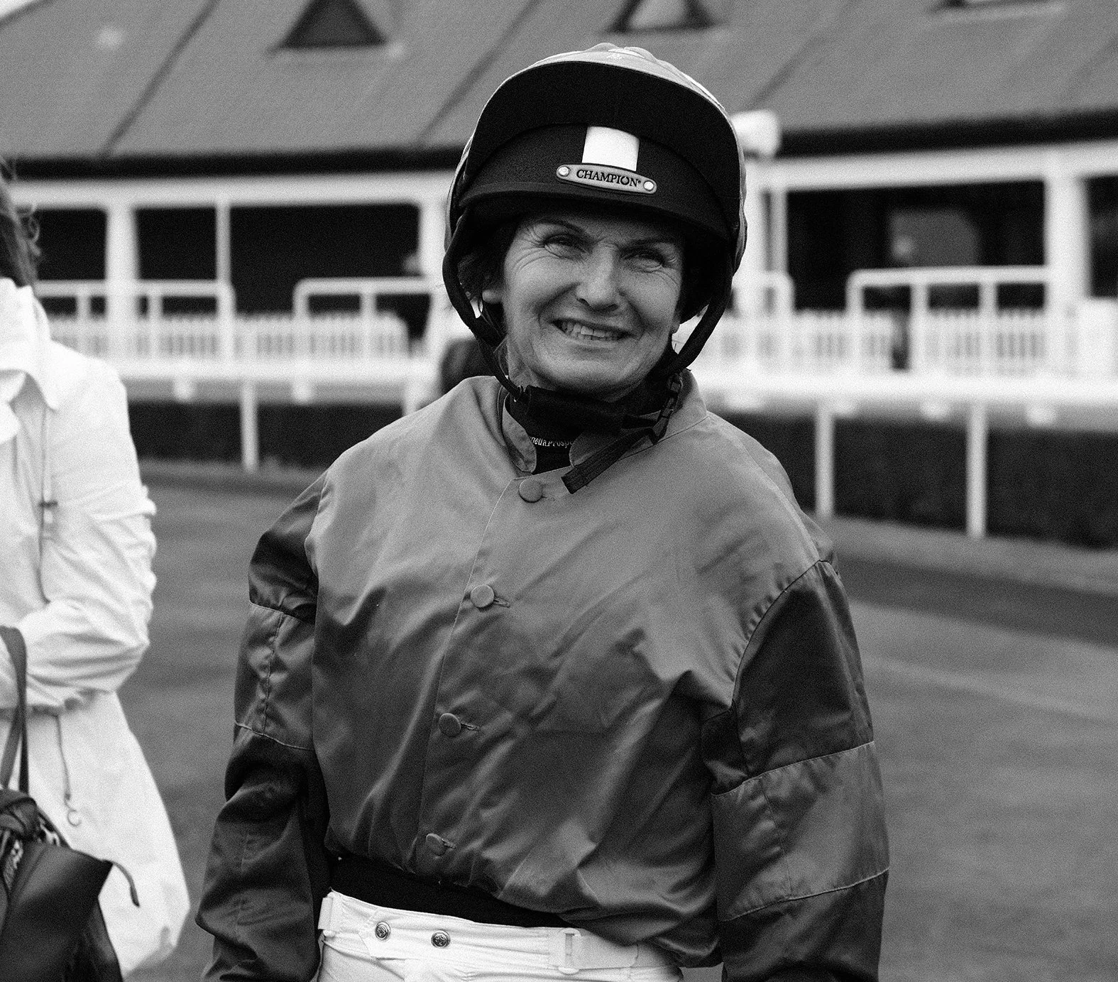 A woman in racing gear smiling, wearing a helmet with the word 'Champion' on it, at a racetrack.