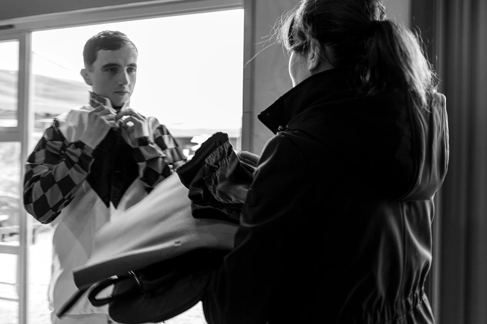A young male race car driver adjusts his racing suit while a woman hands him a helmet in a garage or pit area.