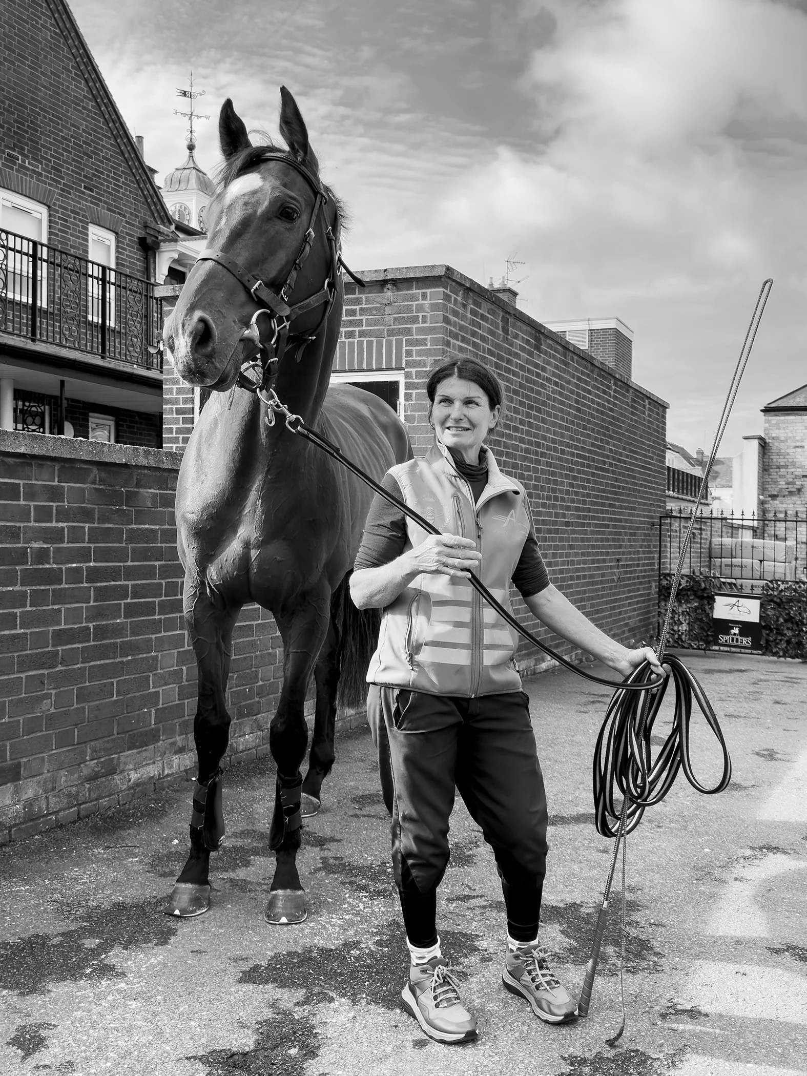 A woman with short dark hair, wearing a sleeveless vest, holding a lunge whip and leash, standing next to a tall horse in an urban alleyway with brick buildings and cloudy sky in the background.