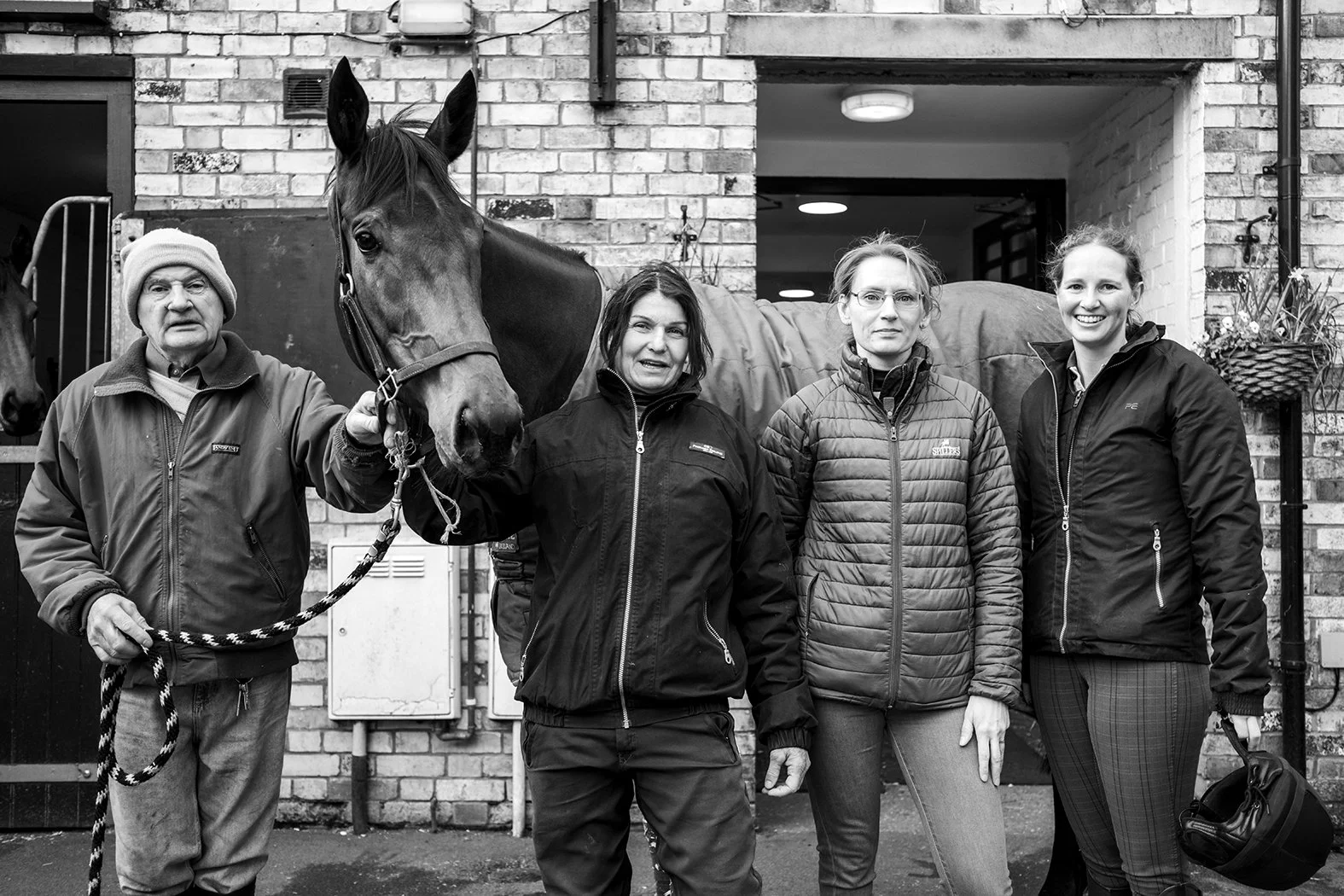 A black and white photo of four women and an older man standing outside in front of a brick building, with a horse in the middle. The man holds the horse's bridle, and the women are standing close together, all dressed in jackets. The woman on the fa