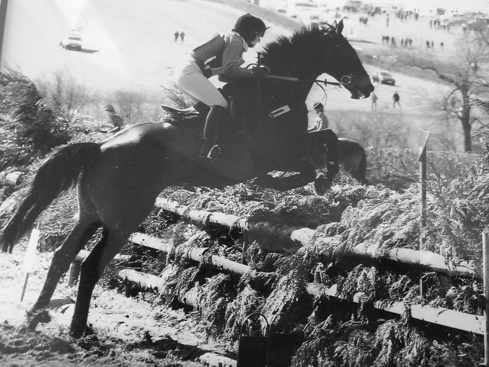 Black and white photo of a horseback rider jumping over an obstacle during an equestrian event, with other spectators and horses in the background.