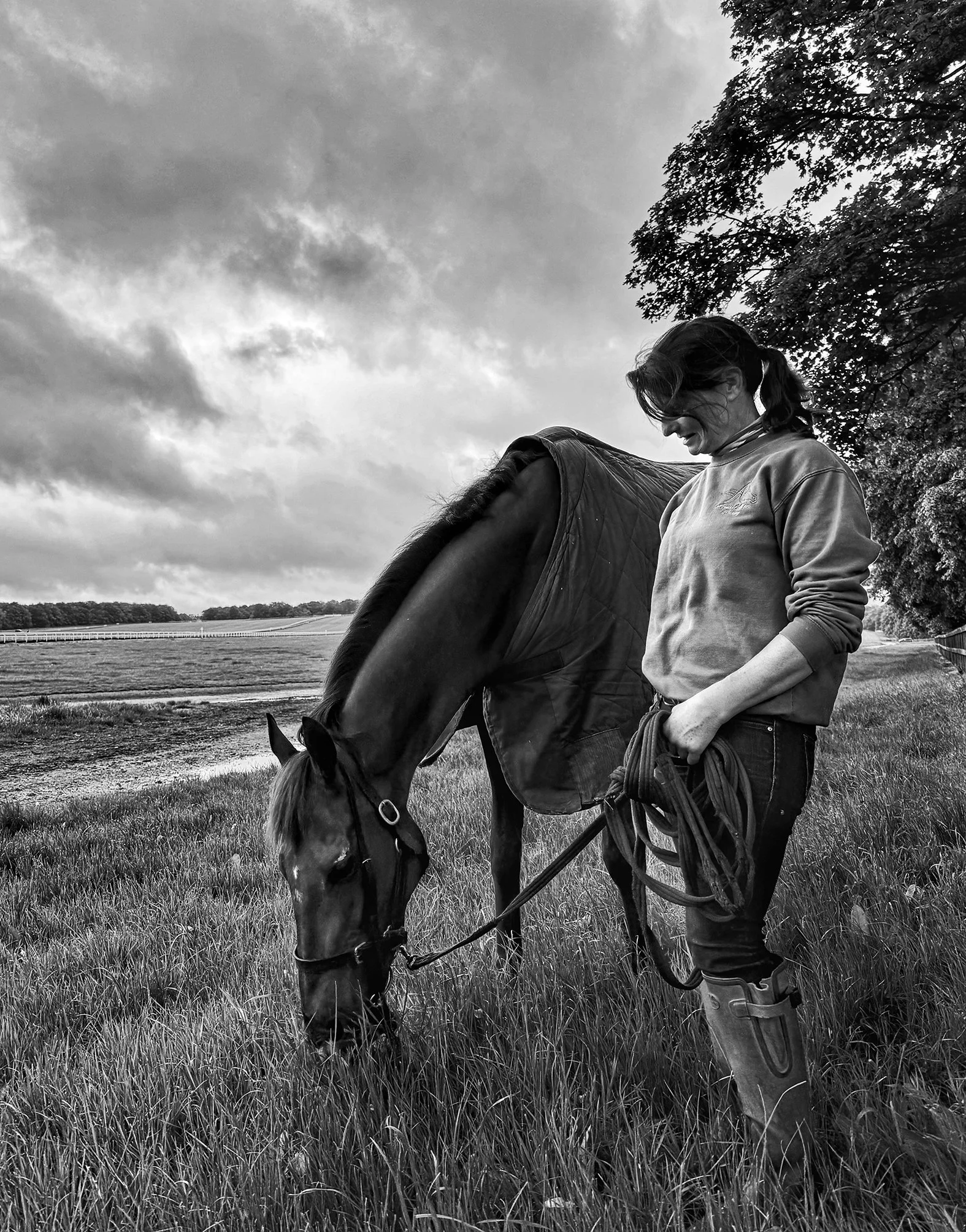 A woman with dark hair tied in a ponytail, wearing a sweatshirt and rubber boots, standing in a grassy field holding a lead rope attached to a horse's bridle, as the horse grazes on the grass. The scene is outdoors with trees and a cloudy sky in the 