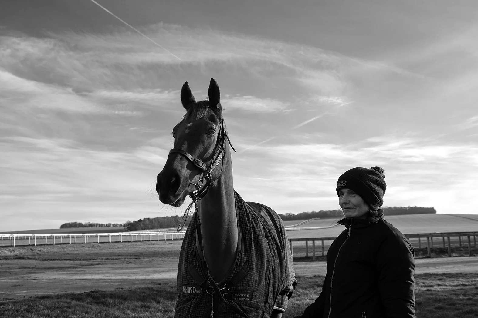 A woman stands next to a racehorse on a racetrack in black and white, with a partly cloudy sky overhead.