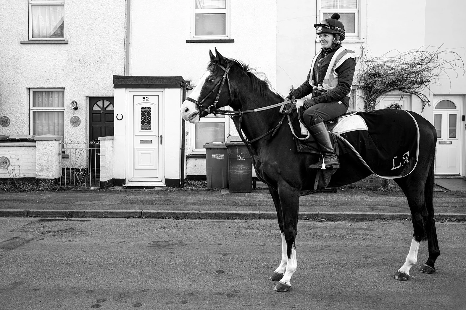 A woman in riding gear, including a helmet and high boots, sitting on a black and white horse on a residential street. The background features a row of houses with front doors and windows.