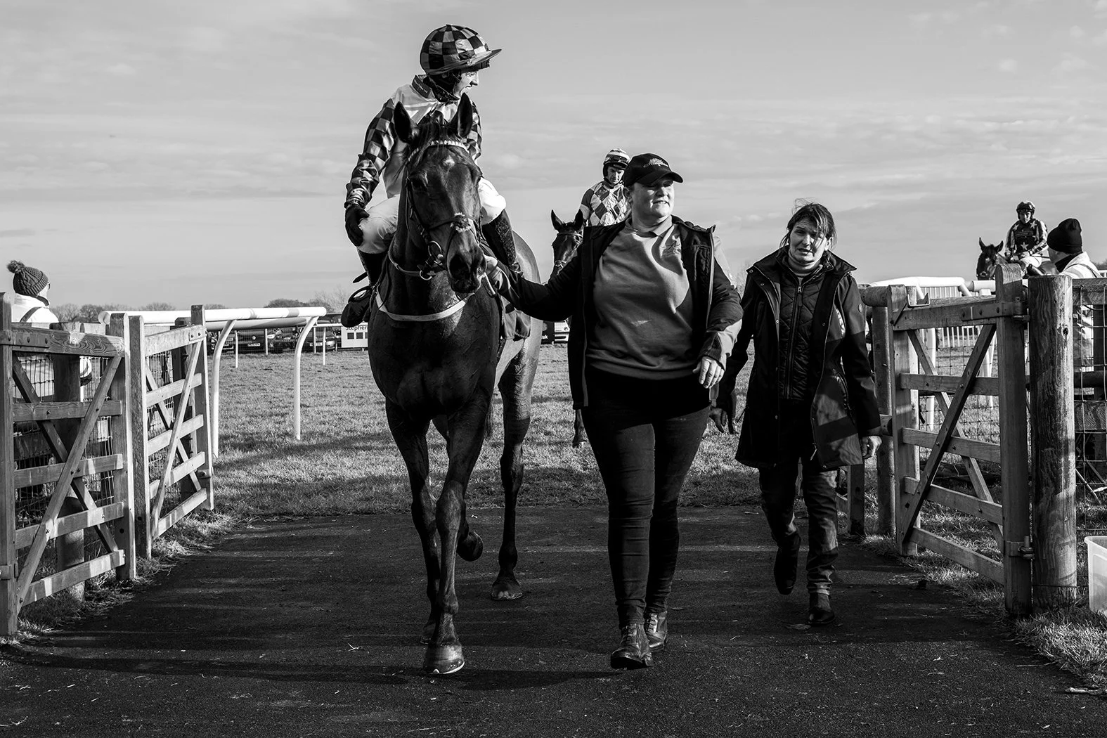 Black and white photo of a woman leading a horse with a jockey riding it, at a racetrack. The woman is walking on a paved path while the jockey holds the reins. Other jockeys and horses are visible in the background near the fence.