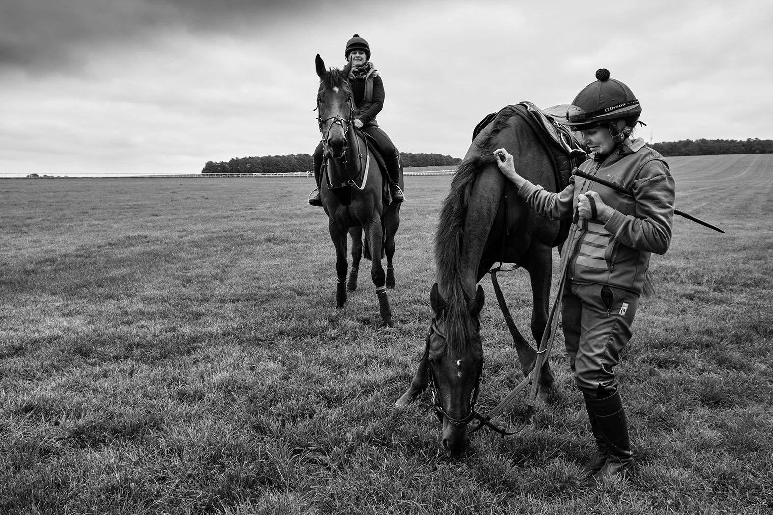 A woman on horseback and a man standing beside a foal, on a grassy field with a cloudy sky in the background.