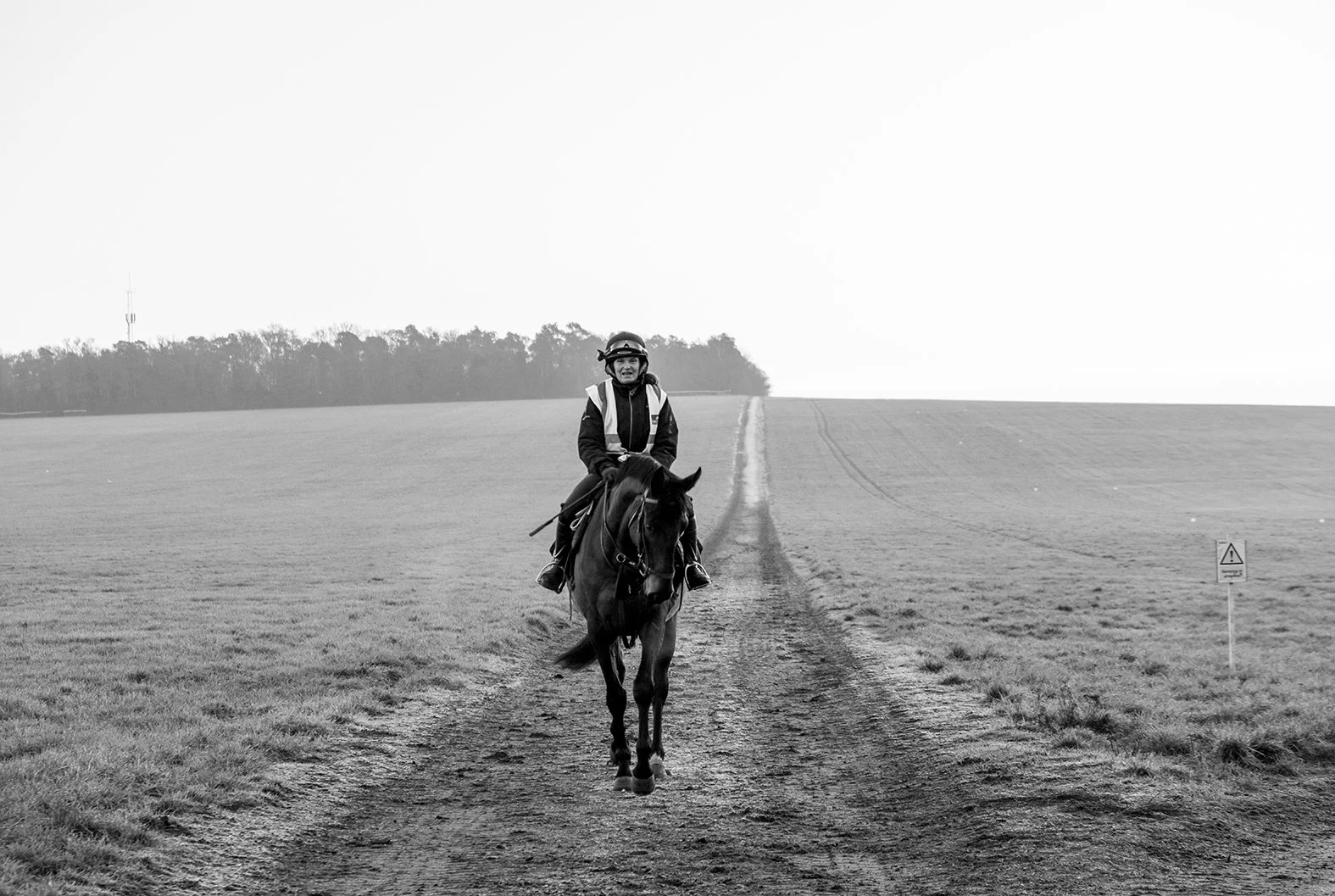 A person wearing a helmet and a safety vest riding a horse on a dirt path through an open field, with a warning sign visible on the right side.