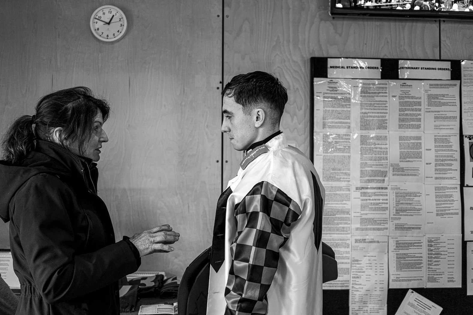 An elderly woman and a young man in a hospital or clinic setting, engaging in conversation, with informational posters on the wall behind them.