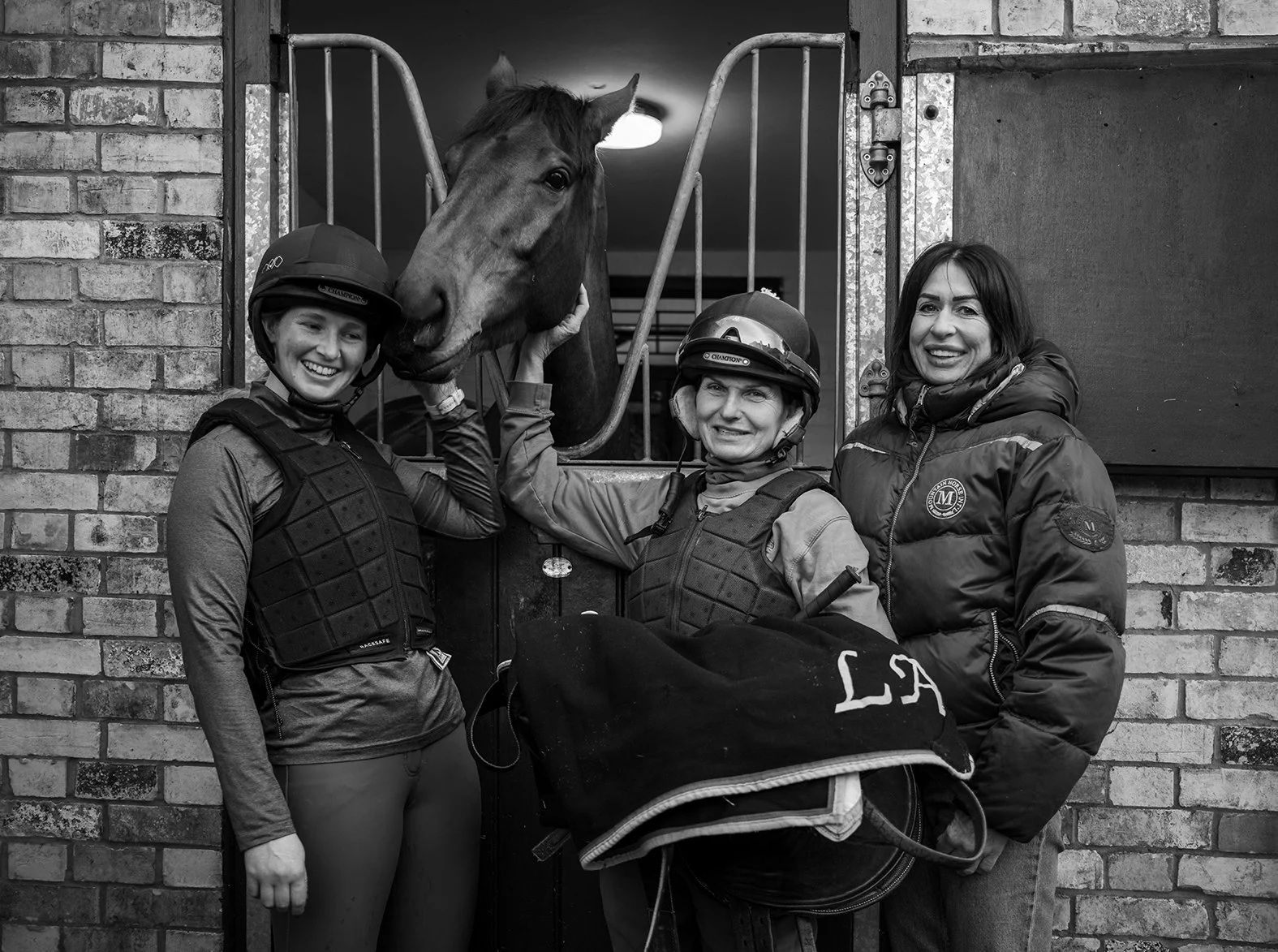 Three women in riding gear and helmets standing outside a stable, with a horse in a stall behind them. One woman is petting the horse, and an equestrian blanket is draped over a saddle.