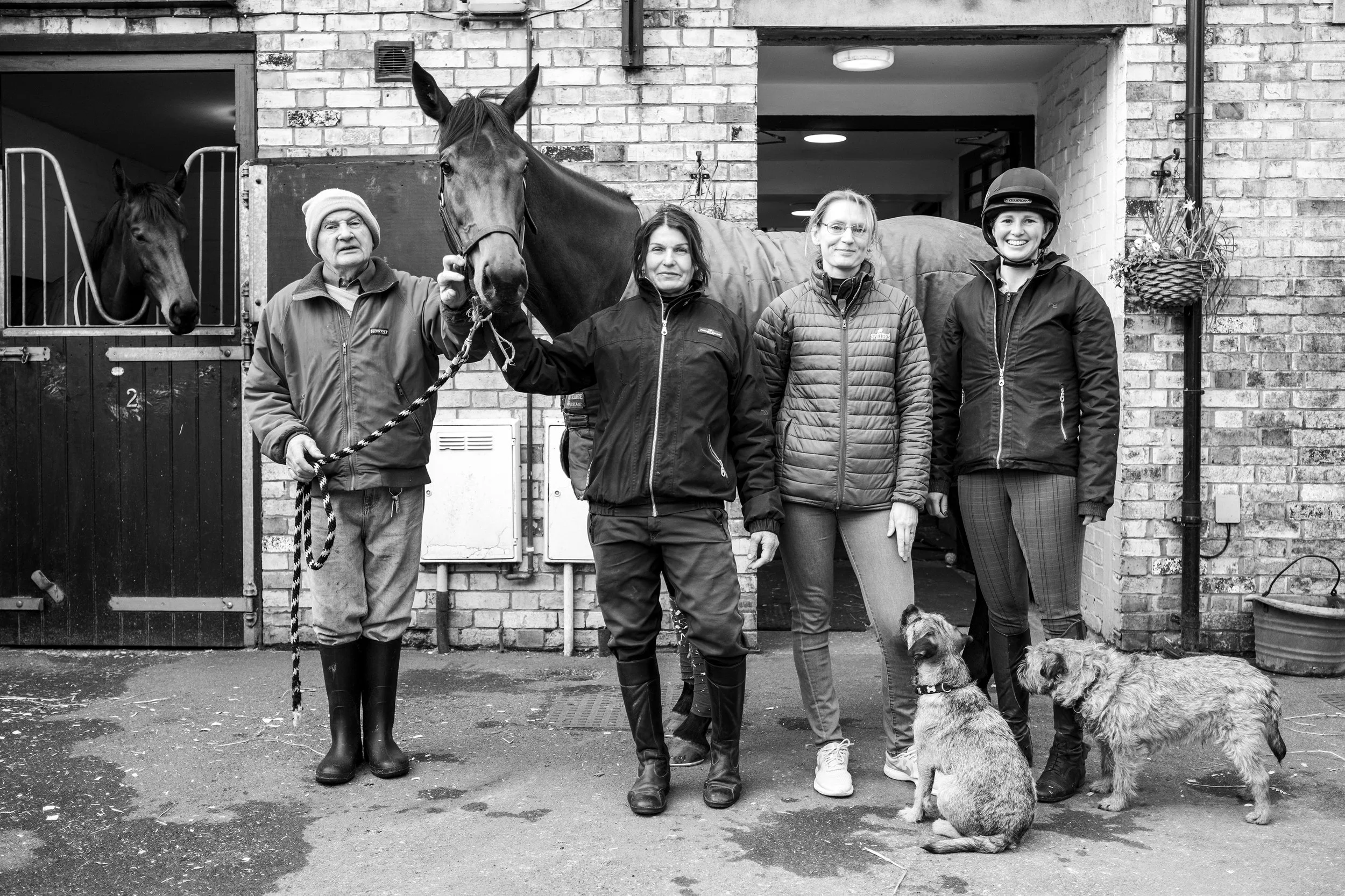 Four people and two dogs standing outside a brick building with a door and a horse stable. One man holds a horse by a lead, and one person wears a riding helmet. Two dogs sit on the ground.