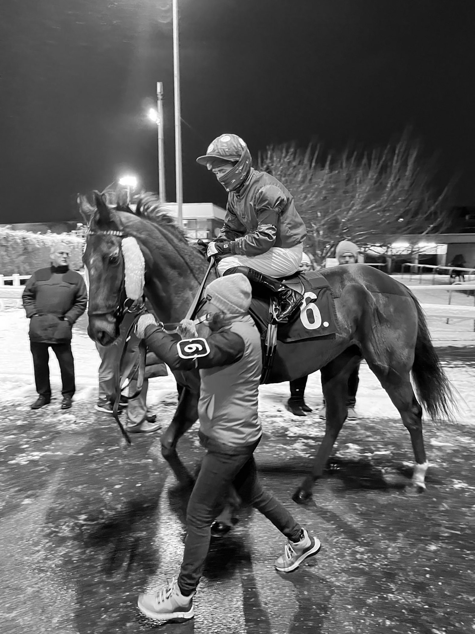 A jockey in riding gear on a horse, with a person helping to secure the saddle, in a nighttime outdoor setting with onlookers and bright lights.