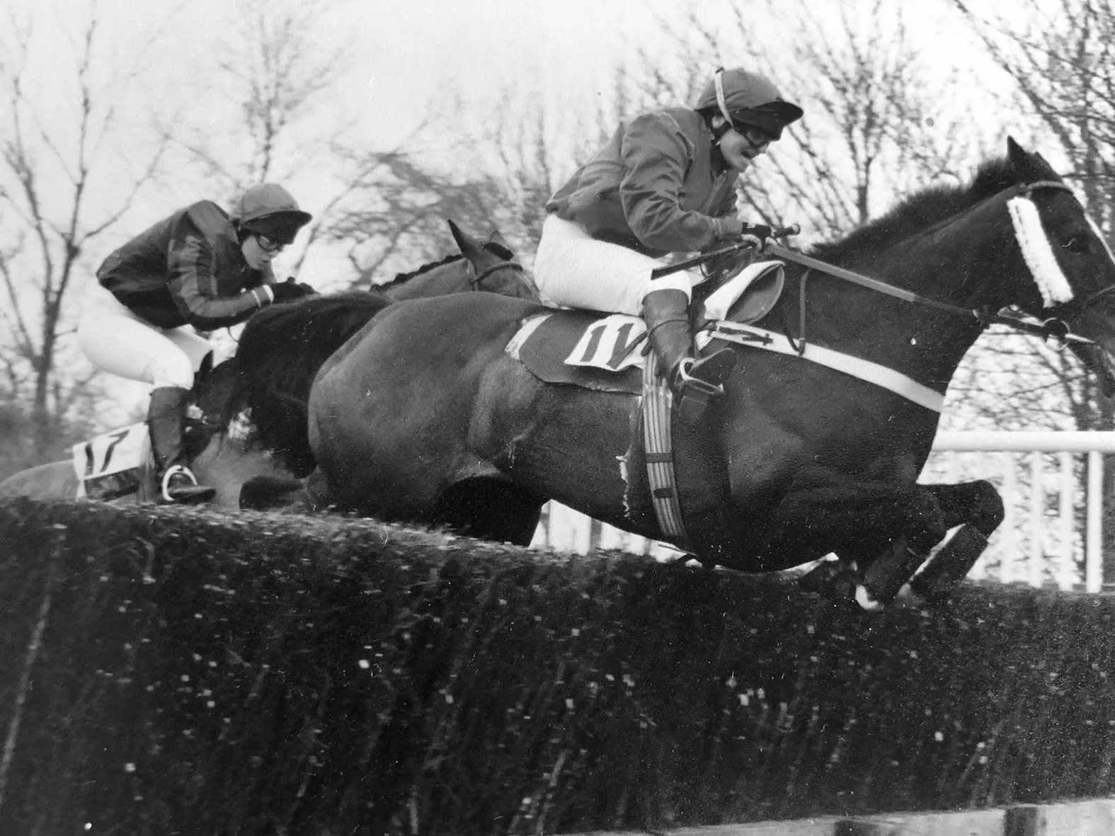 Black and white photo of two jockeys riding racehorses, jumping over a hurdle during a horse race, with trees in the background.