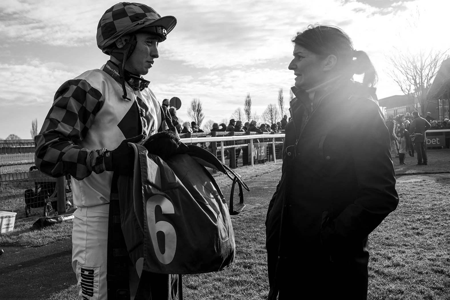 A jockey holding a bag and a woman in conversation at a horse racing track with spectators in the background.
