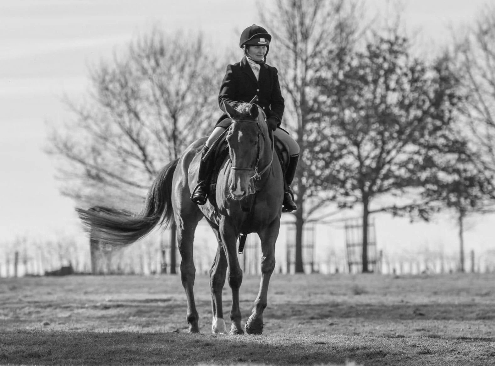 A woman riding a horse outdoors in a park with trees in the background, captured in black and white.