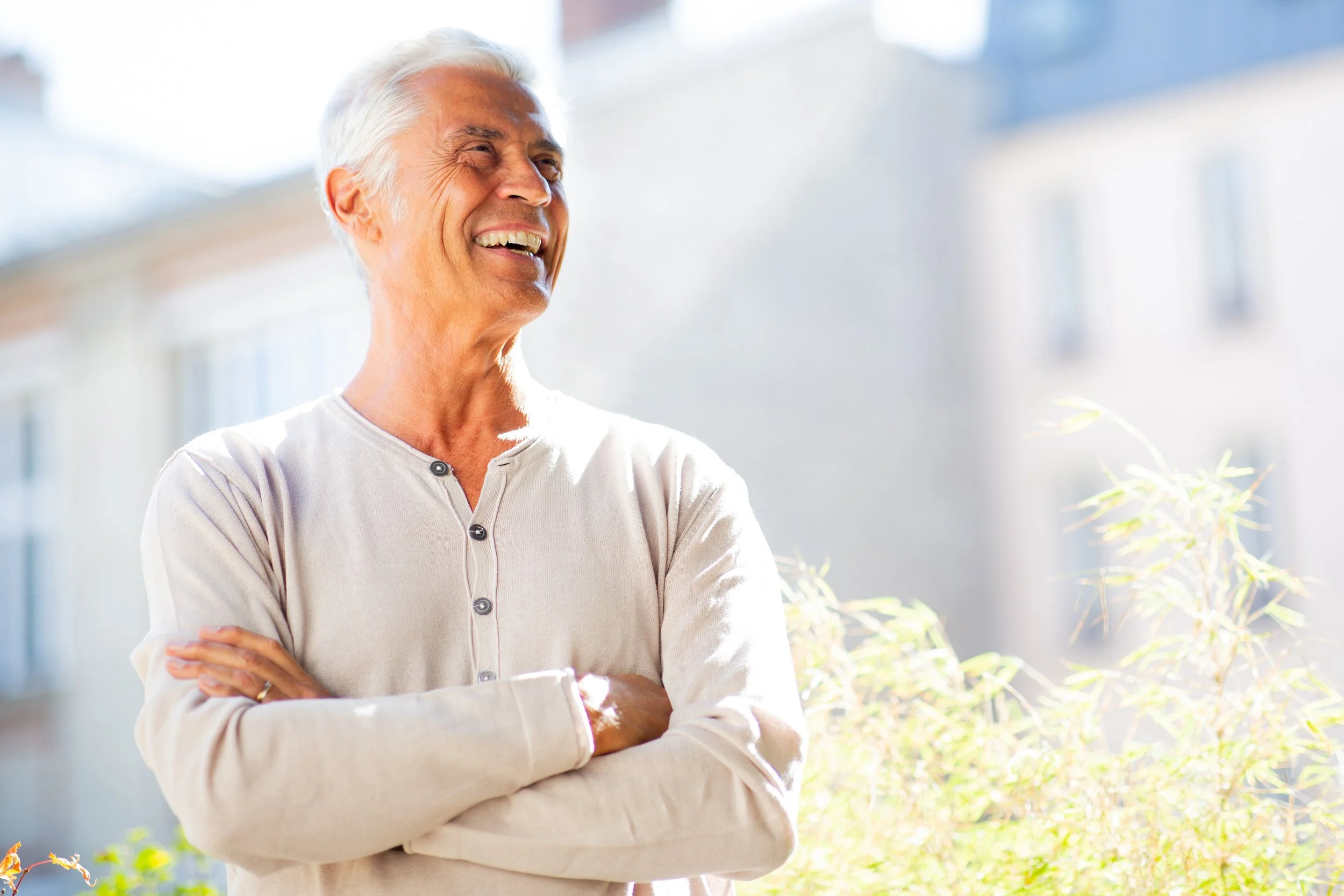 An older woman with gray hair smiling outdoors with arms crossed, bright sunlight, modern buildings in the background.