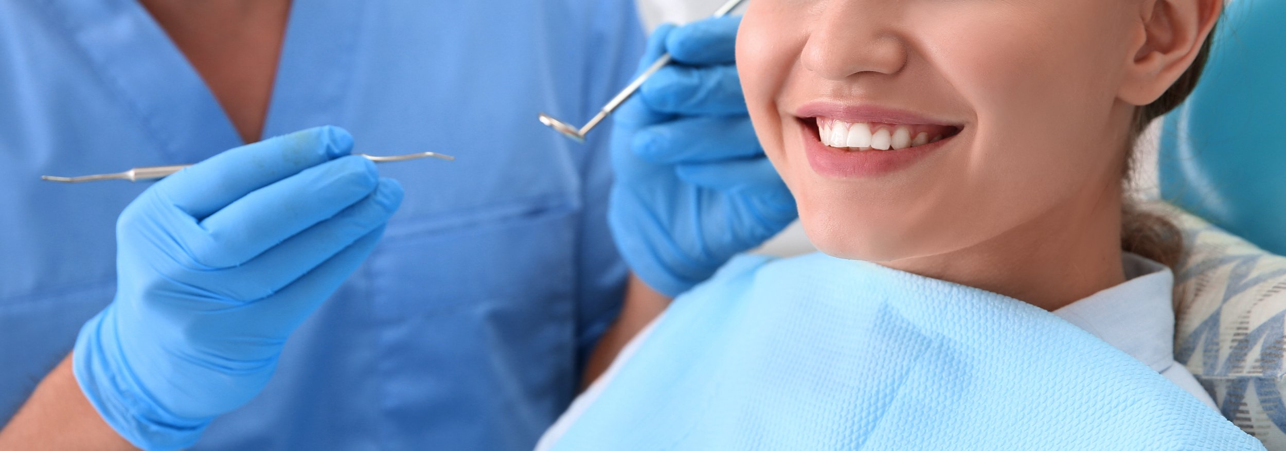 A woman at the dentist's office smiling as a dentist wearing blue gloves prepares to clean her teeth with dental tools.