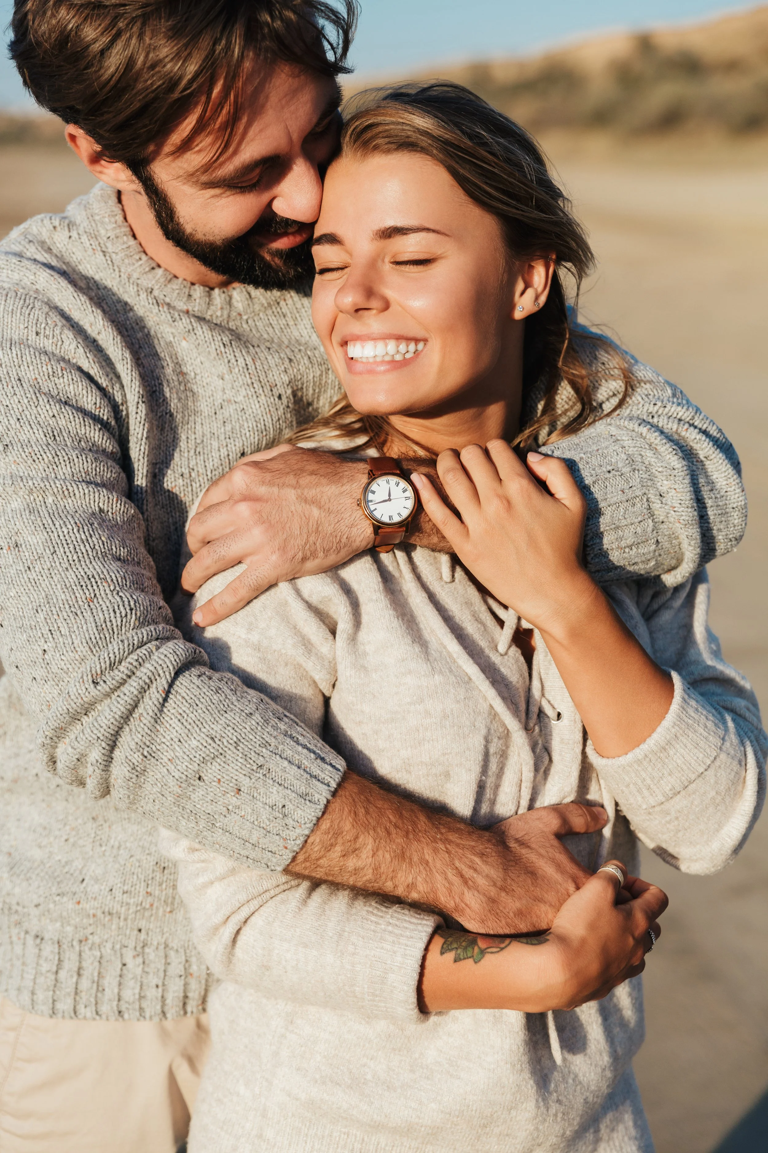 A happy couple with beautiful teeth embracing on a beach during sunset, smiling with eyes closed.