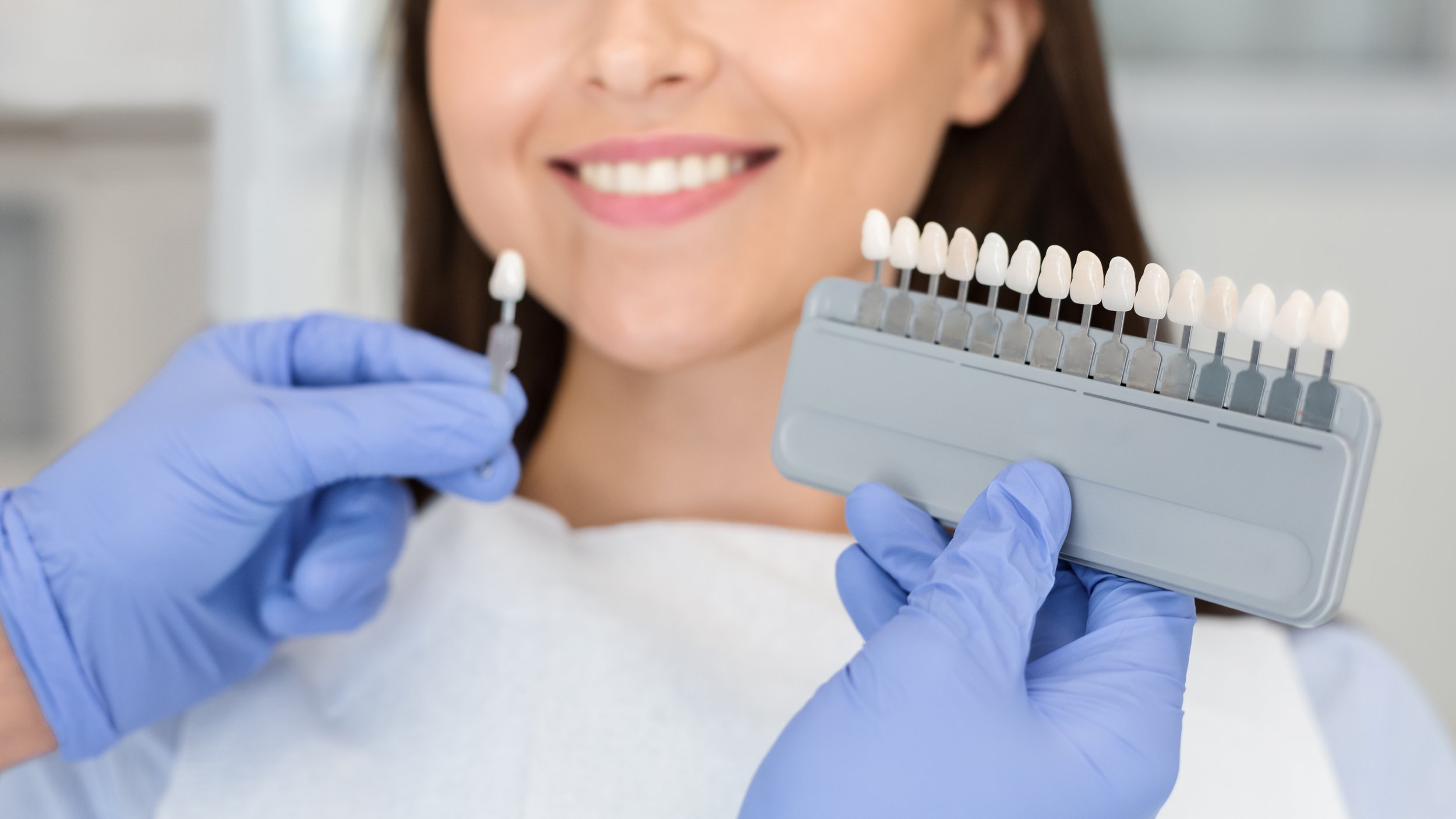 A dentist wearing blue gloves holds a dental shade guide near a patient's mouth, helping her choose the right teeth whitening treatment.
