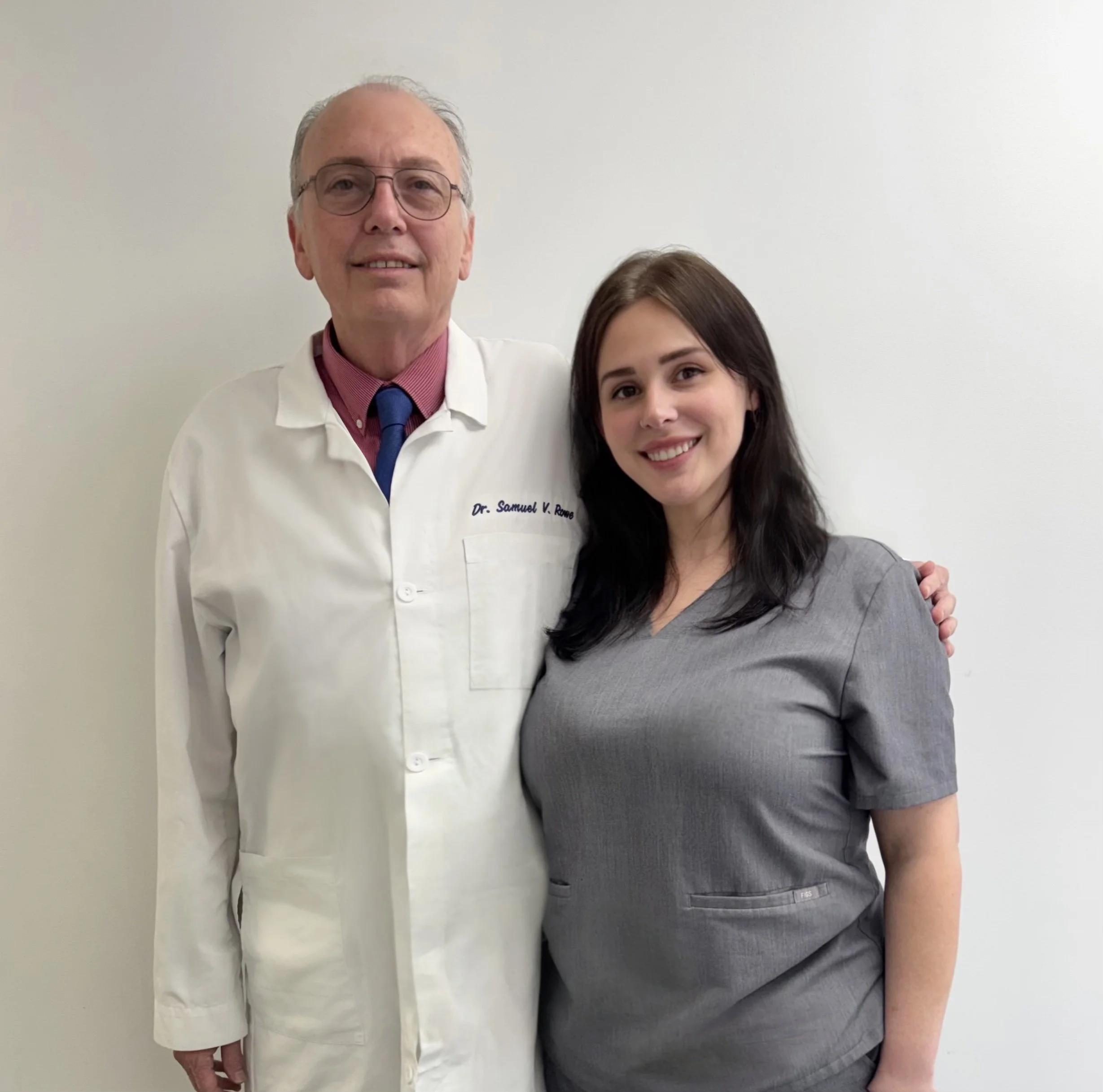 A doctor and a young woman smiling and standing close together against a plain white wall.