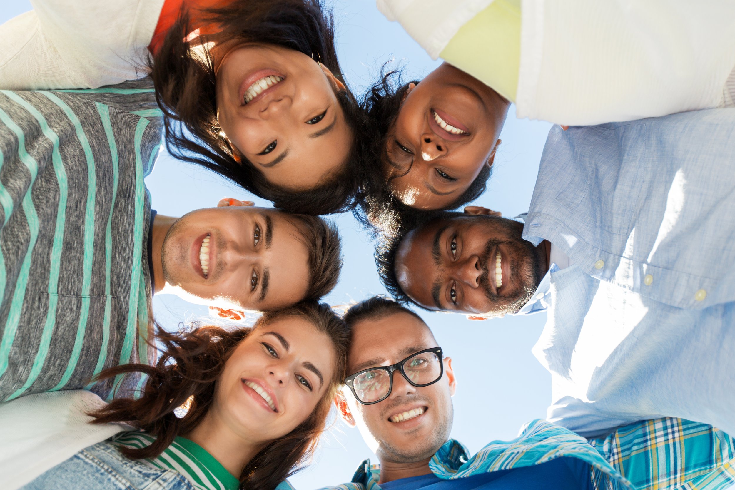 A group of seven diverse young adults smiling and looking down at the camera, forming a circle with their heads outdoors against a clear blue sky.