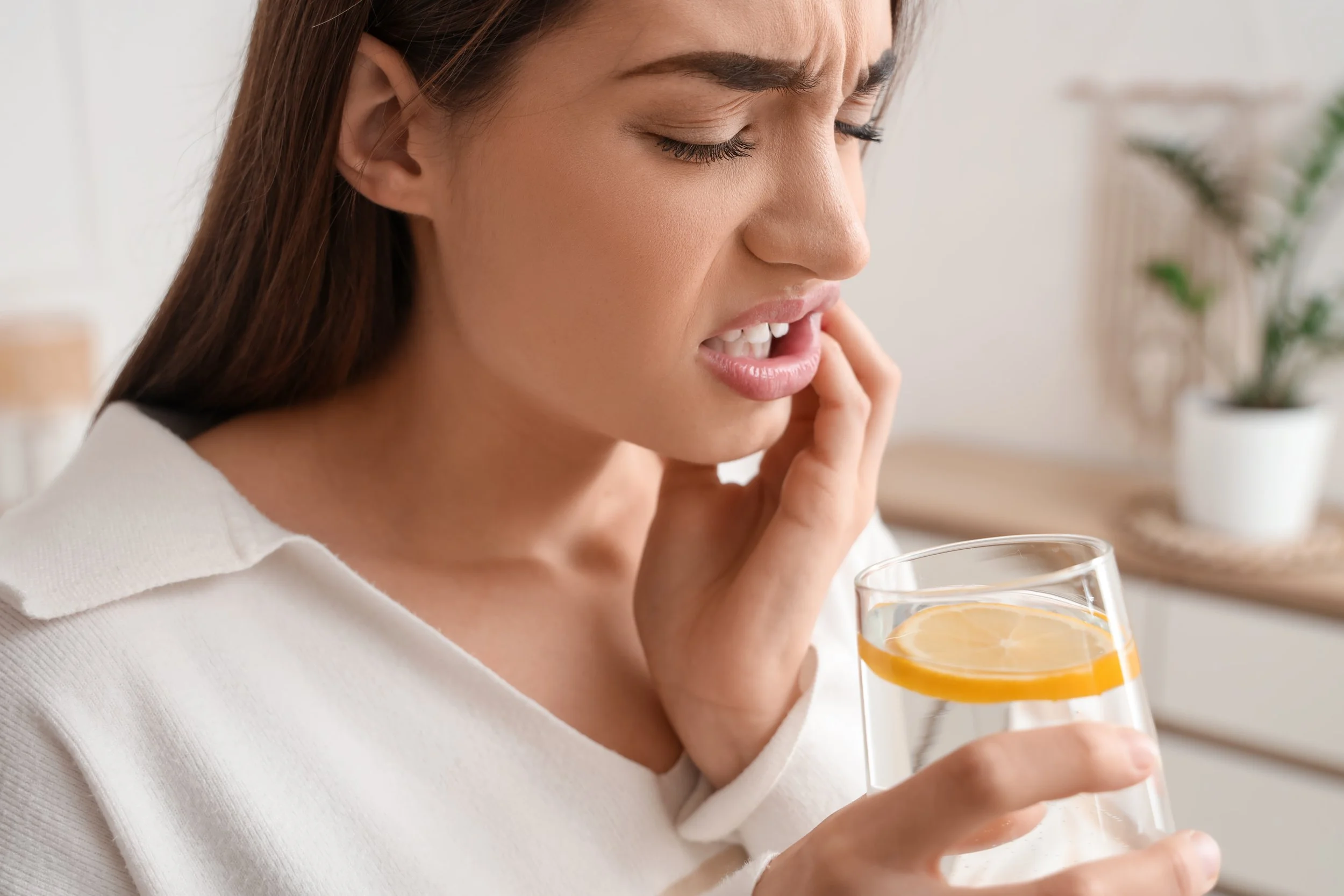A woman with long brown hair holding a glass of water in one hand and her cheek with the other because of tooth pain