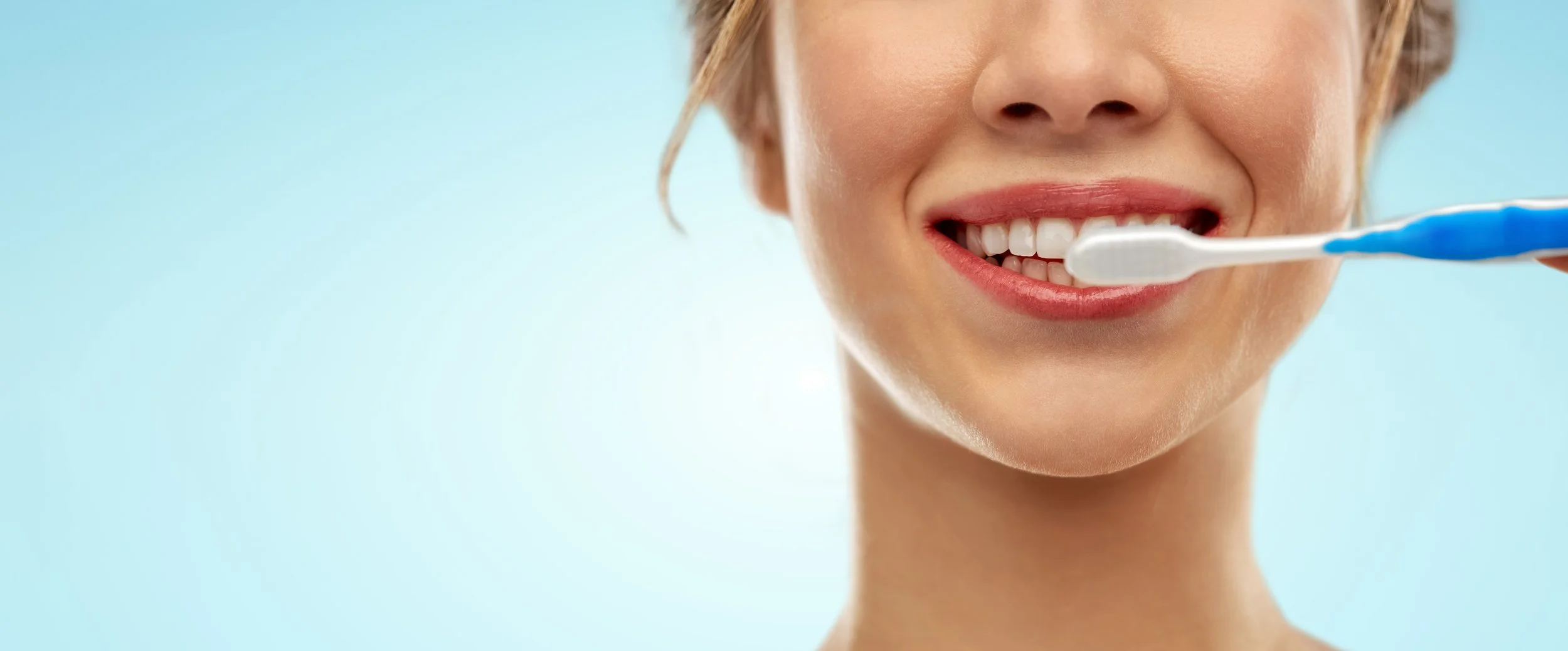 Close-up of a woman implementing dental hygiene with a blue and white toothbrush, smiling in front of a light blue background.