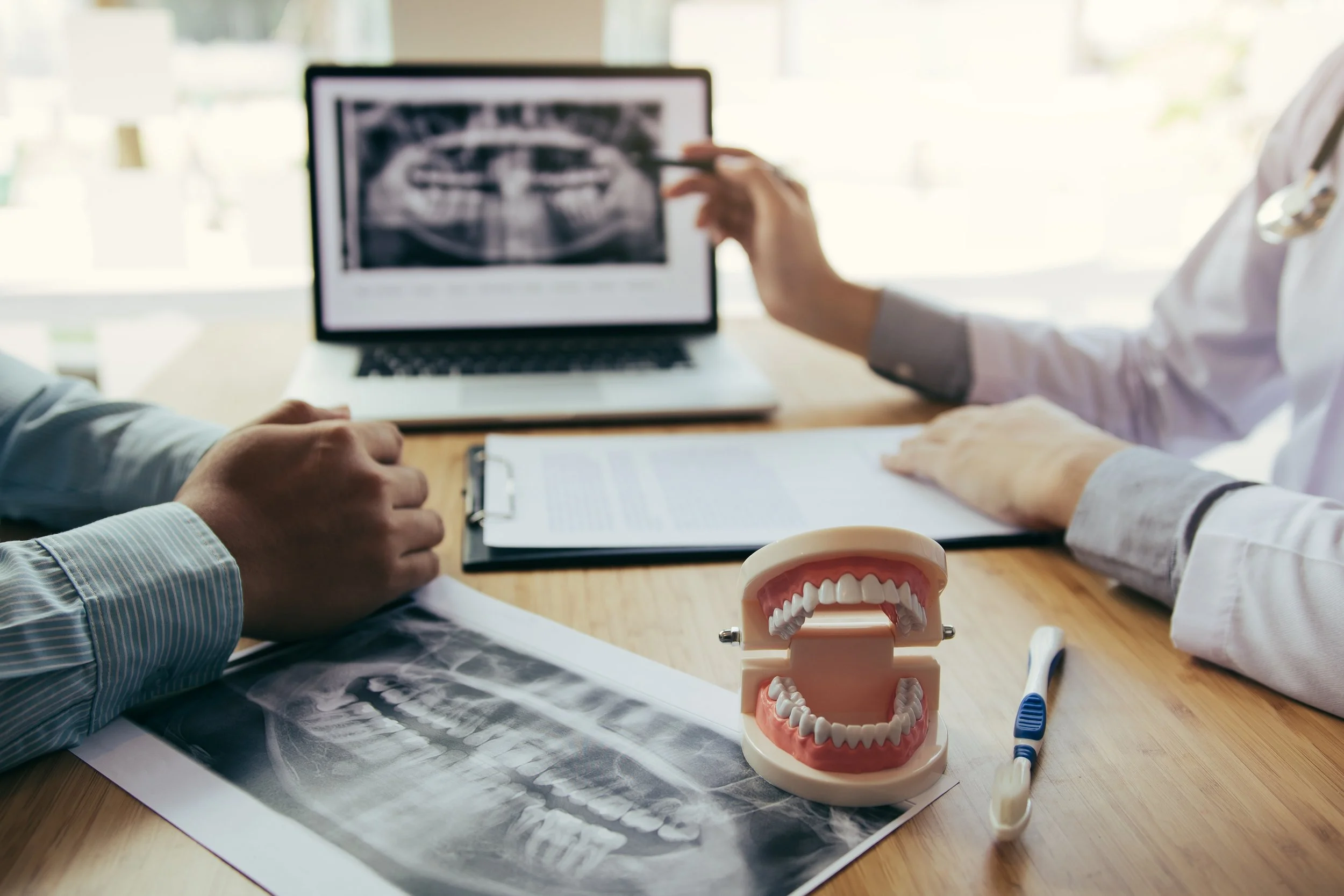 Dental consultation room with a dental model, toothbrush, and dental X-ray on a wooden table, and a dentist and patient discussing a dental scan on a laptop.