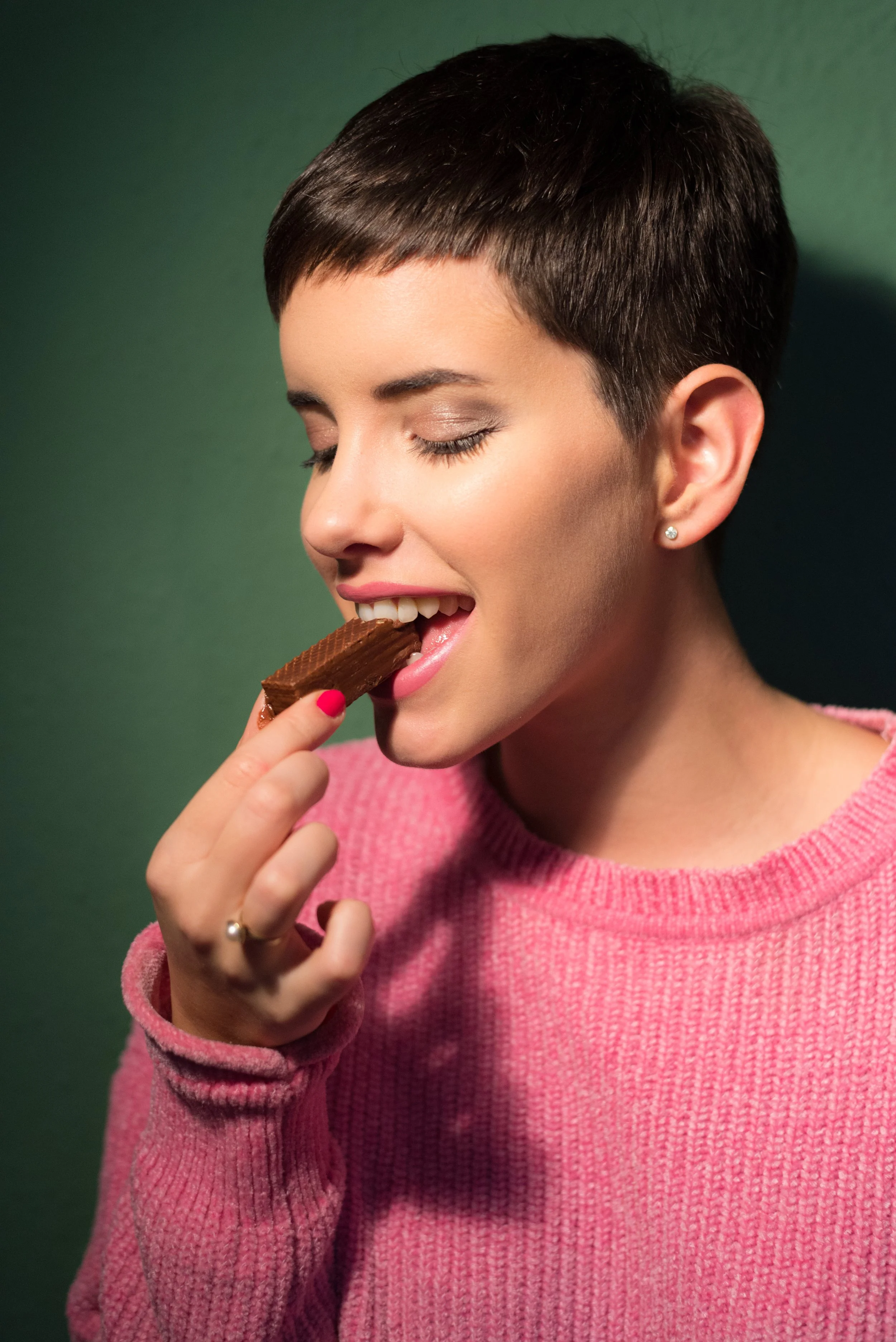 A woman with short dark hair and earrings, wearing a pink sweater, is about to eat a chocolate-covered biscuit, with a green background.