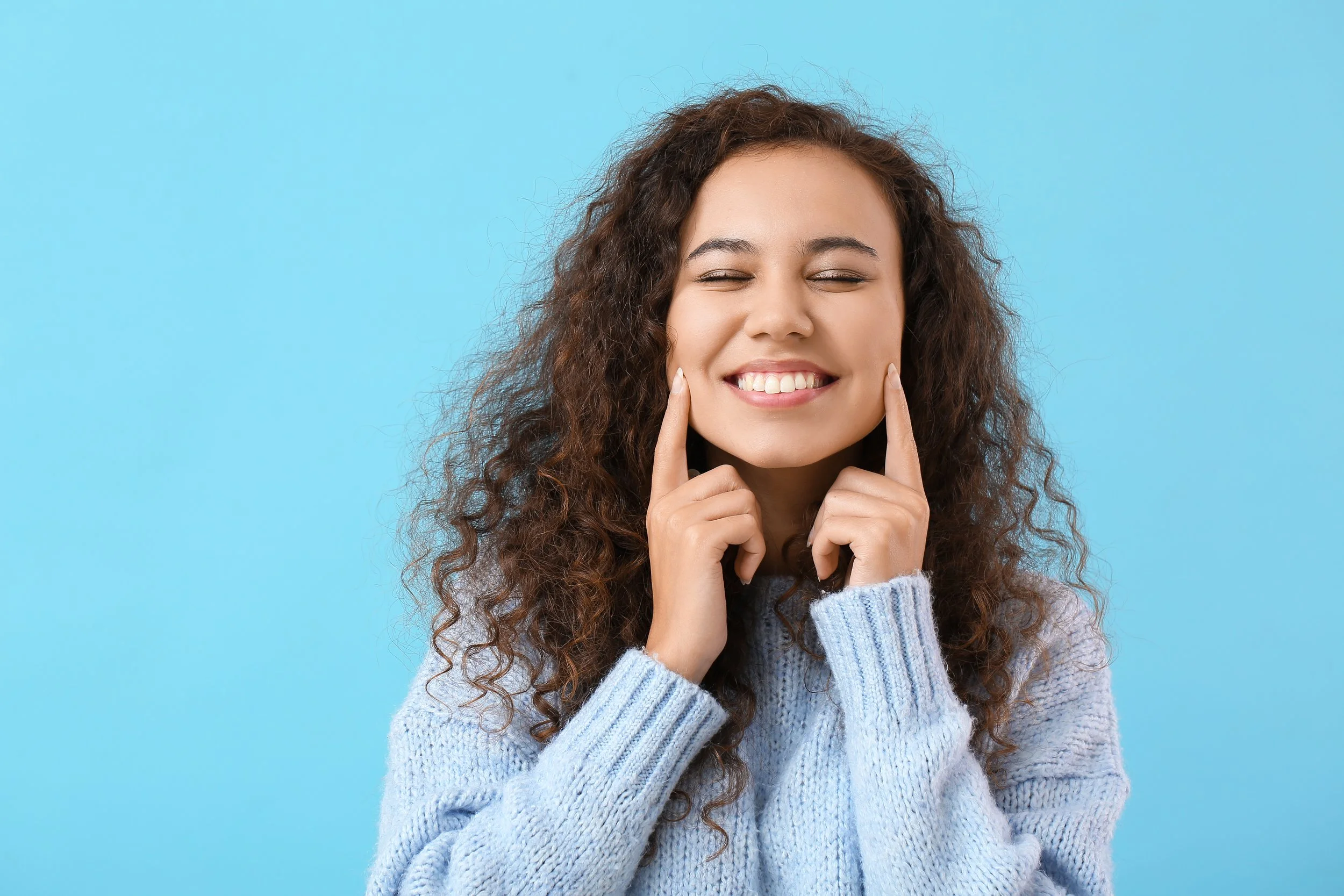 A happy woman with curly brown hair smiling with eyes closed, wearing a light blue knitted sweater against a light blue background. Pointing at her cheeks to show off her tooth bonding.