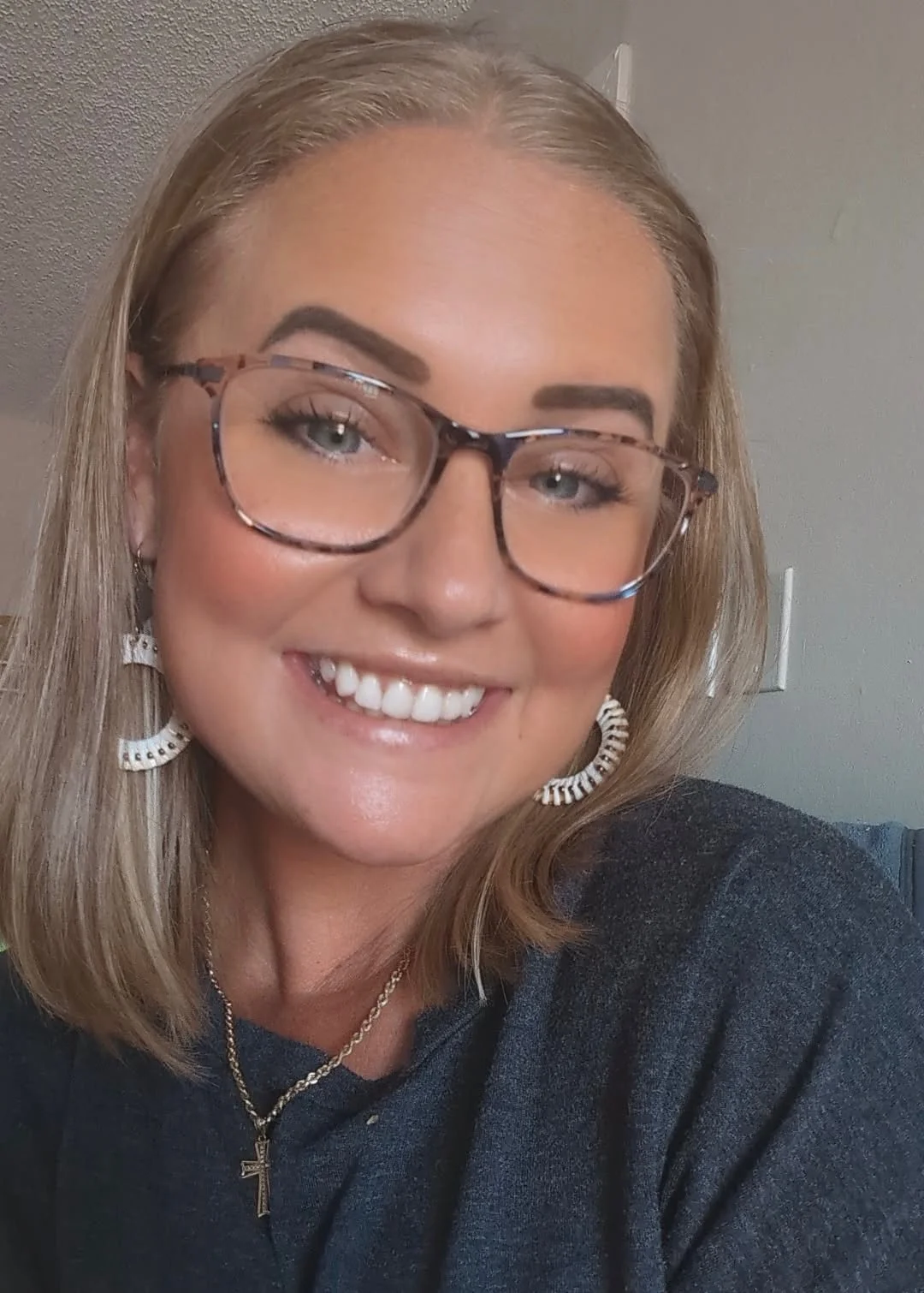 Dental assistant, Angela, a smiling woman with blonde hair, wearing glasses, hoop earrings, a gold necklace with a cross, and a dark top.