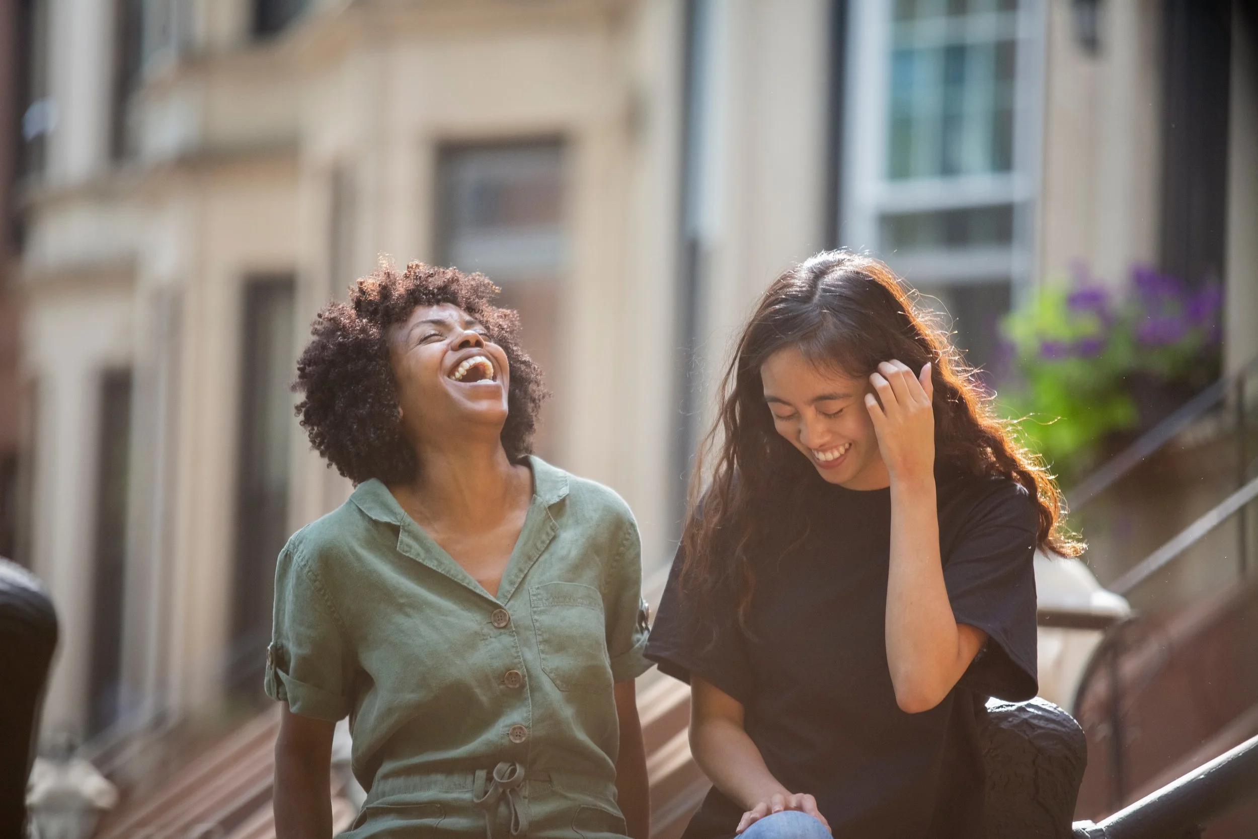 female friends laughing while sitting in front of houses in the city