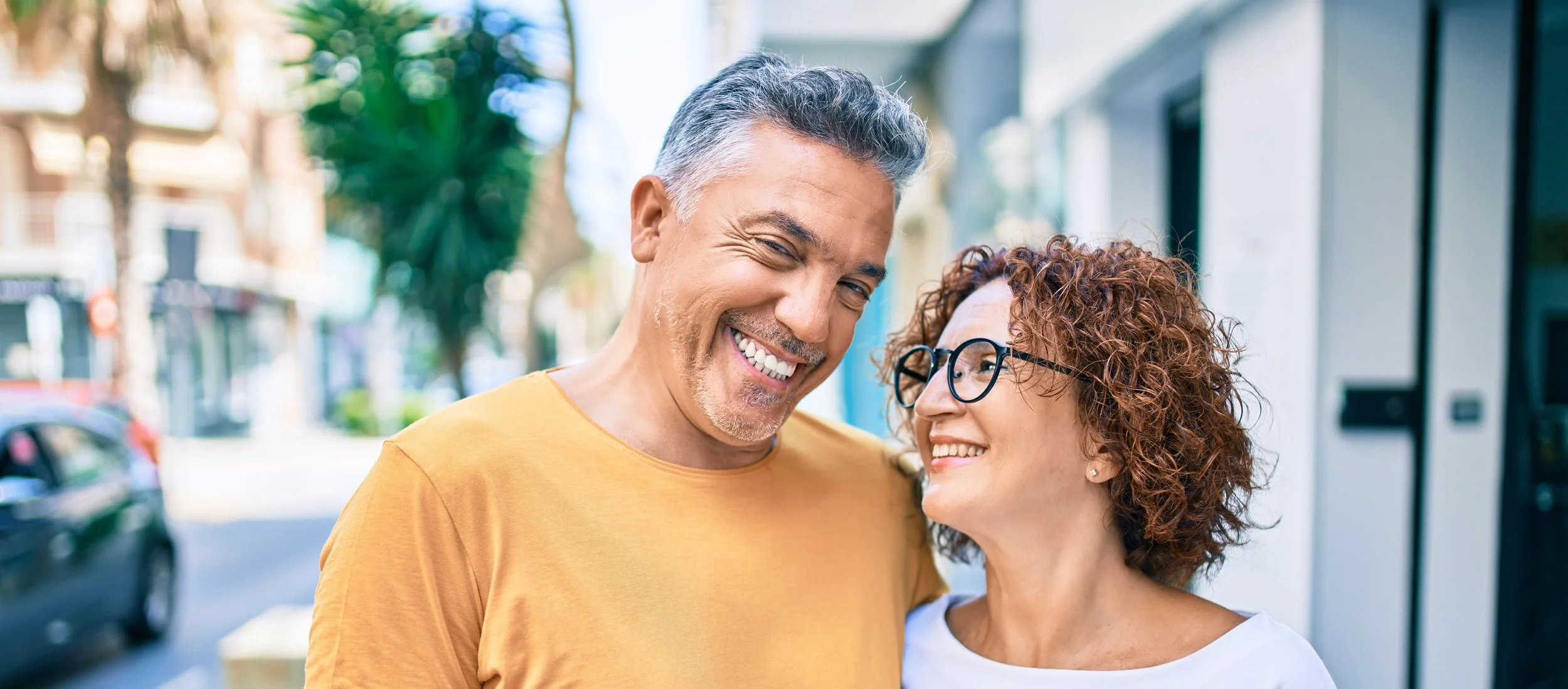 A smiling middle-aged man and woman sharing a joyful moment. The man has gray hair and a beard, wearing a yellow t-shirt. The woman has curly red hair and glasses, wearing a white shirt. Both are laughing with their new dentures.