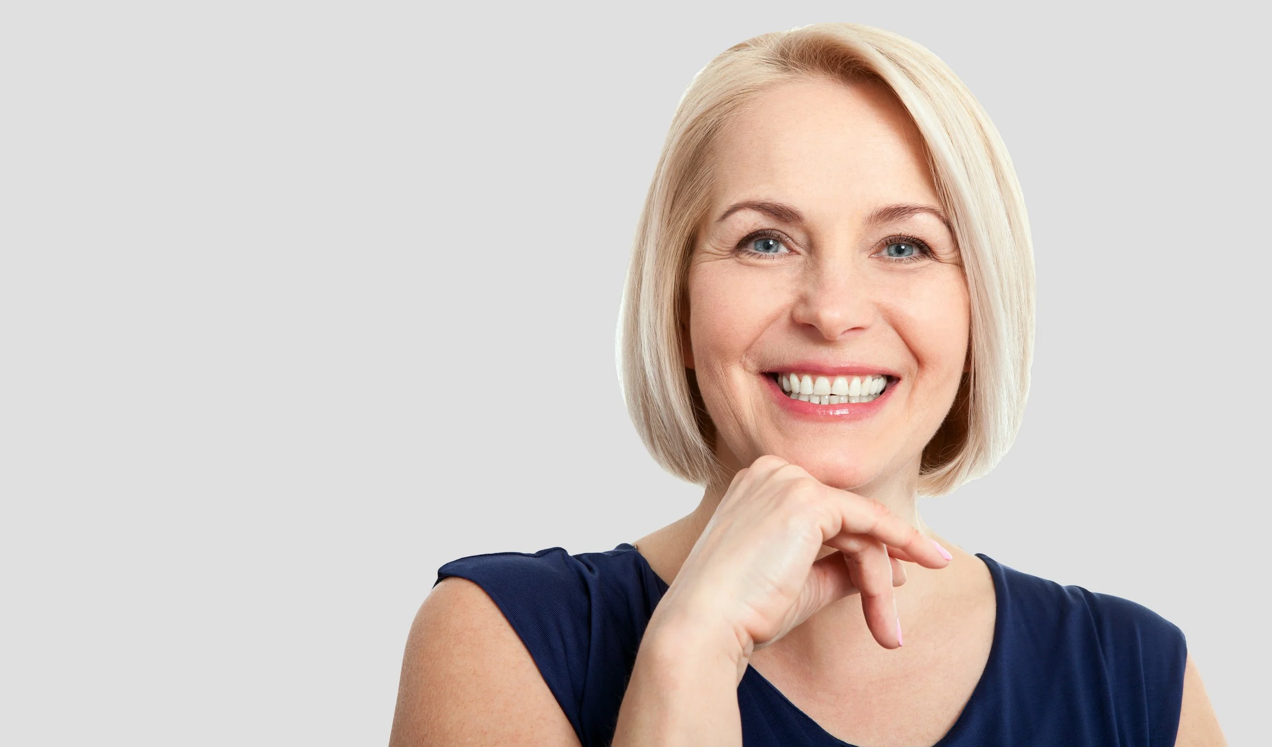 A smiling woman with blonde hair, wearing a navy blue sleeveless top, posing with her hand near her chin.